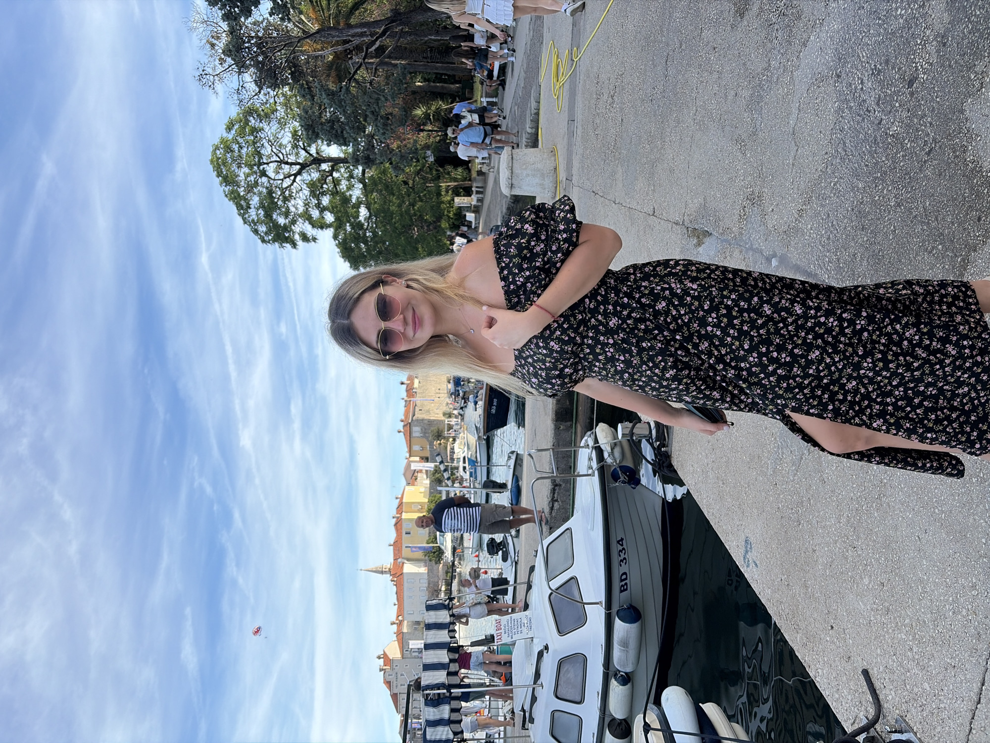 A young woman with blonde hair and sunglasses stands on a concrete pier in Budva, Montenegro. She is wearing a black off-the-shoulder dress with a floral pattern and a high slit. Behind her, a fleet of small boats is docked in the harbor, with a picturesque town featuring red-tiled roofs and a church spire visible in the background. The sky is blue with scattered clouds, and a parasailer can be seen in the distance. Other people are walking along the pier and in the background, suggesting a lively tourist area. The overall scene is a sunny day at a coastal European town.