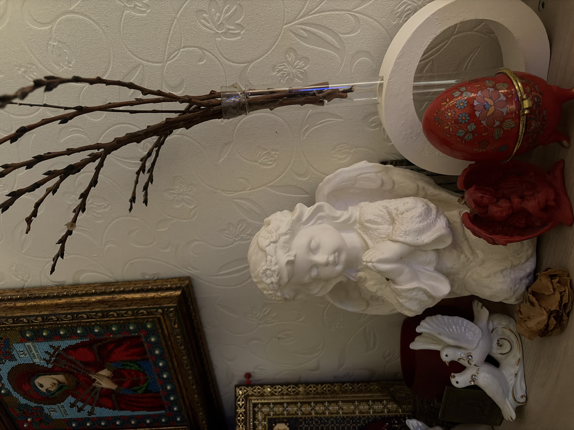 The image shows a collection of objects arranged on a surface, possibly a shelf. These objects include a small, white angel figurine, a vase with bare branches, a decorative red egg-shaped object, a white circular frame, a framed image of a religious figure, a red object that might be a candle holder, and a white porcelain dove figurine. The background is a textured white wall. It is not possible to confirm the location of Chaikovsky, Russia, based on the image alone.