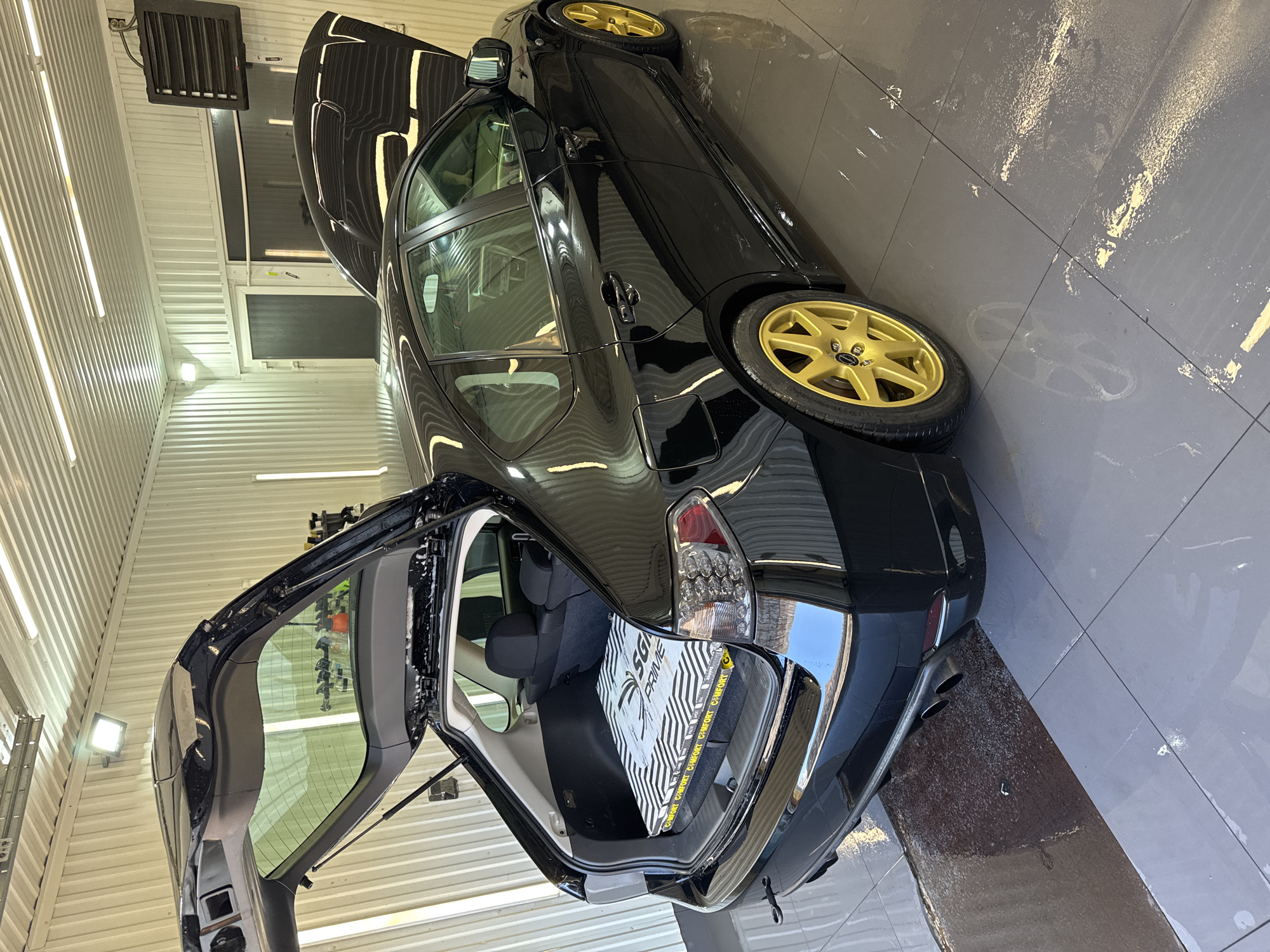 The image shows a black Subaru hatchback with its hood and trunk open, parked inside a well-lit garage or detailing shop. The car has gold-colored wheels. There's a box in the trunk with "SGI PRIME" visible on its side. The floor appears to be wet, possibly from washing or detailing. The walls are lined with corrugated metal or paneling. A heater is mounted high on the wall. Based on the query including the city name, it is assumed that the car is in Chaikovsky, Russia.