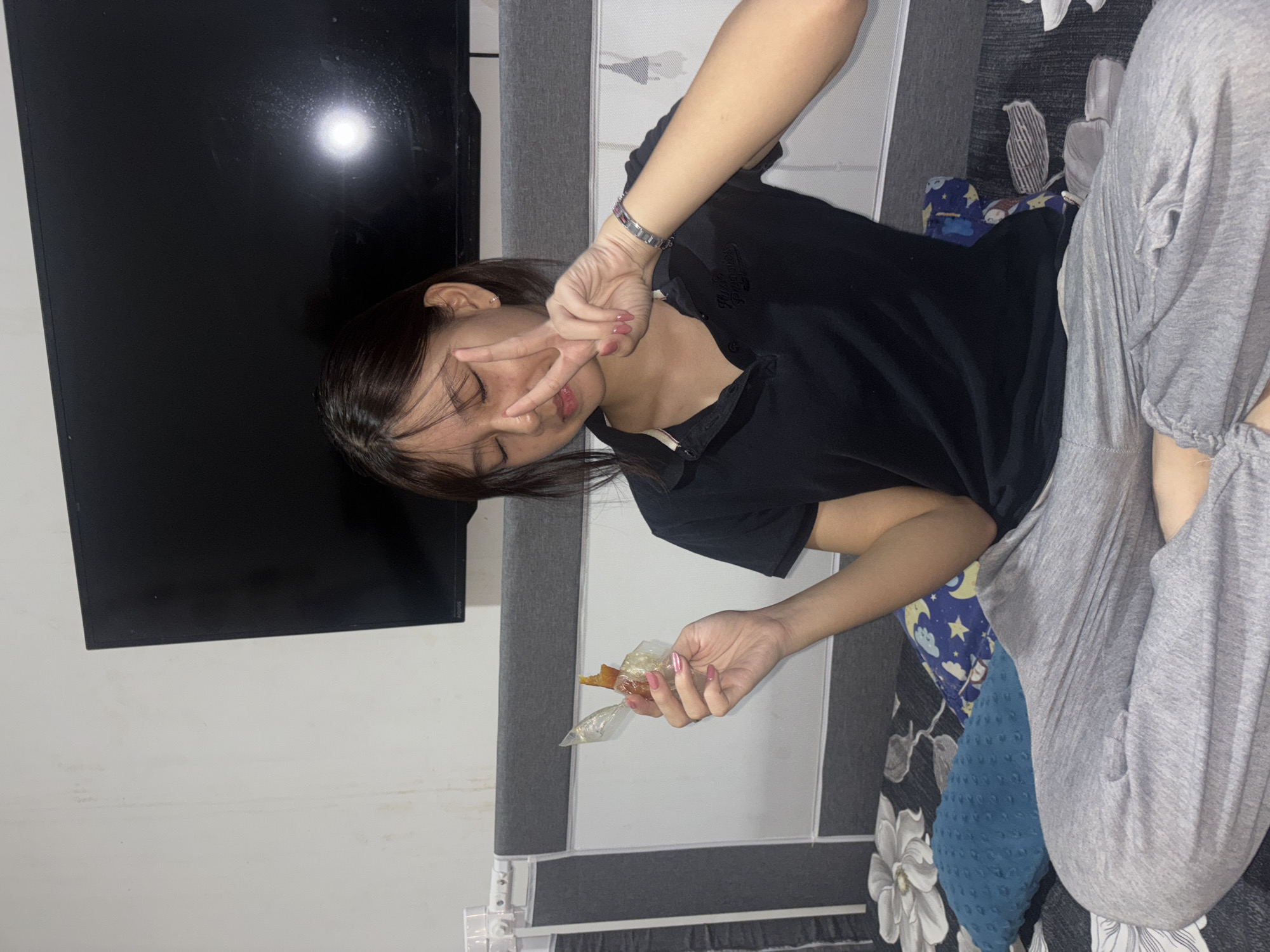 The image depicts a young woman sitting cross-legged on a bed in what appears to be a bedroom. She is wearing a black polo shirt and grey sweatpants. Her eyes are closed, and she is holding a piece of food wrapped in plastic in her left hand, while her right hand is raised in a peace sign. A bracelet is visible on her right wrist. Behind her, a large flat-screen television is mounted on the wall, and the bed is covered with patterned bedding and pillows. The lighting suggests an indoor setting, likely at night. The provided context indicates the location is West Jakarta, Indonesia, implying the scene is within a residential dwelling in that area.