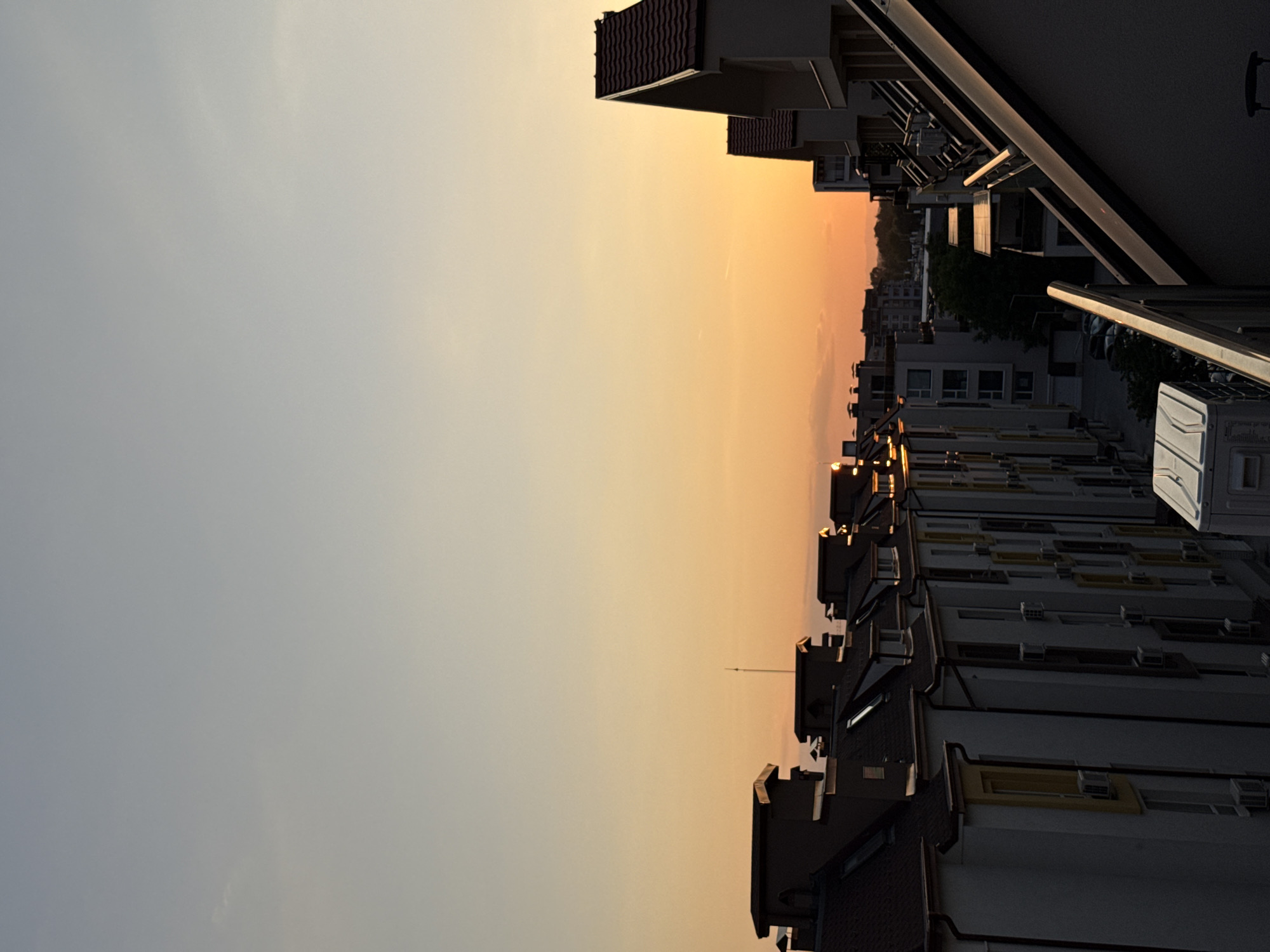 The image shows a cityscape in Plovdiv, Bulgaria, taken during sunset. Rows of buildings with tile roofs and light-colored facades line the frame, receding into the distance. The sky is gradated from grey to orange, indicating the setting sun. Some balconies are visible, and the photo appears to be taken from a high vantage point. The image is oriented sideways.