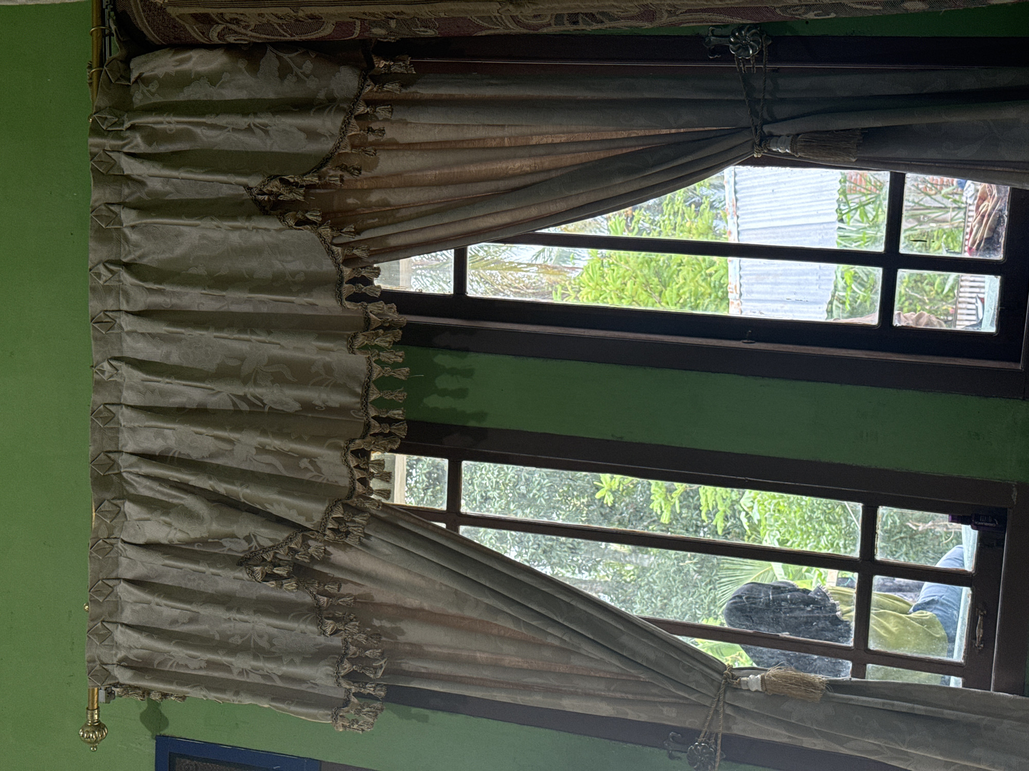 An indoor, eye-level medium shot captures a pair of open curtains with tassels and ornate trim, framing two adjacent windows that look out onto a lush green outdoor environment. The curtains are a muted gold color with a subtle floral pattern and are drawn open by tasseled tie-backs. The window frames are dark brown wood. The left window shows a person wearing a black hijab and a lime green garment, partially visible through the glass. The right window offers a view of dense foliage and a corrugated metal roof. The wall surrounding the windows is painted a vibrant green. The lighting suggests it is daytime.