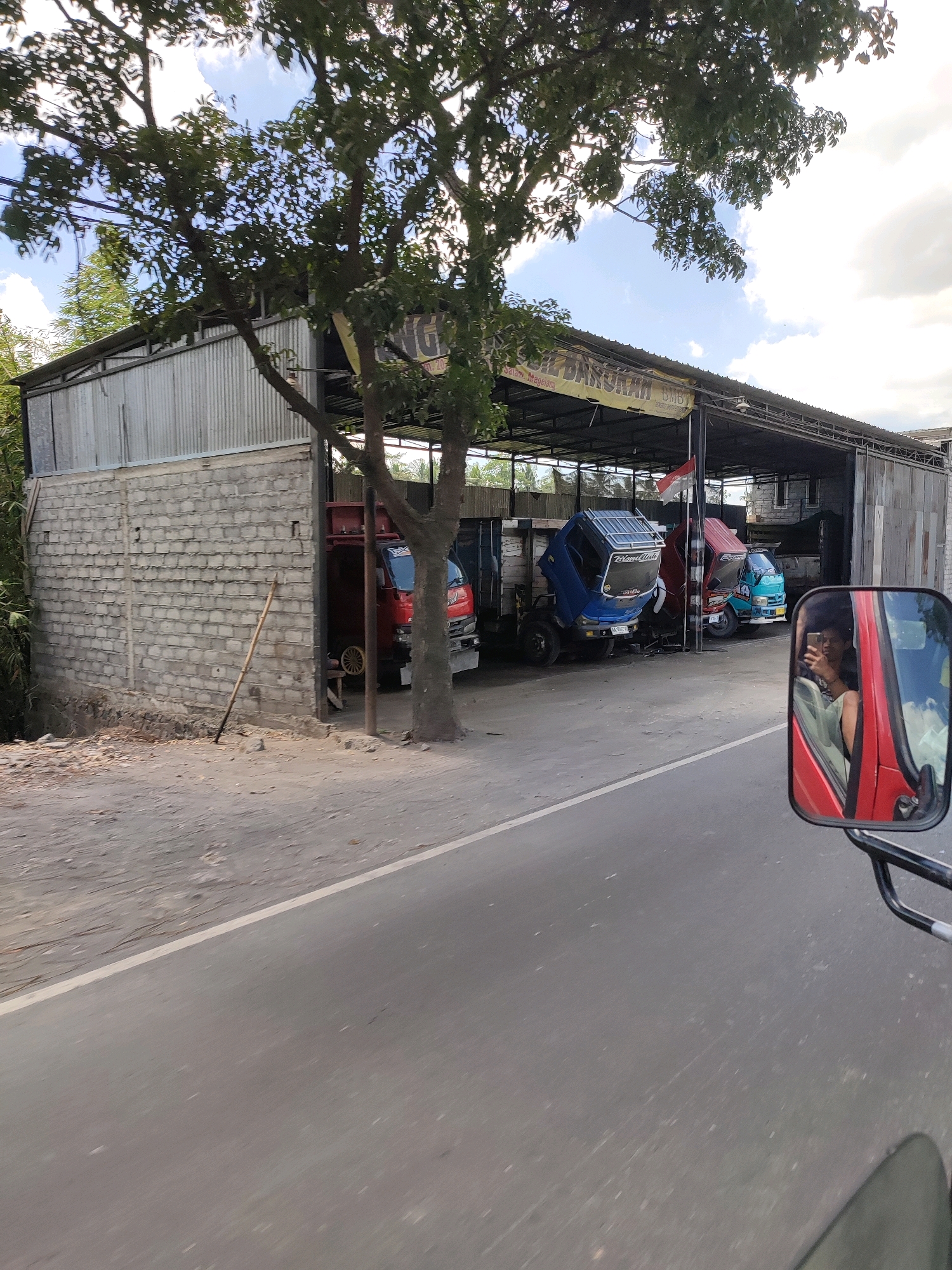 A side view of a garage with three trucks parked inside is captured from a moving vehicle's side mirror. The garage has a corrugated metal roof supported by metal poles, with a brick wall on the left side. A large tree with green leaves partially obscures the view of the garage and sky. The garage appears to be located on the side of a road in Salam, Indonesia, as indicated by the text on a banner above the garage that reads "BENGKEL BMB Salam-Magelang". In the side mirror of the vehicle, a young man is seen taking a selfie. The road in front of the garage is paved and dusty.