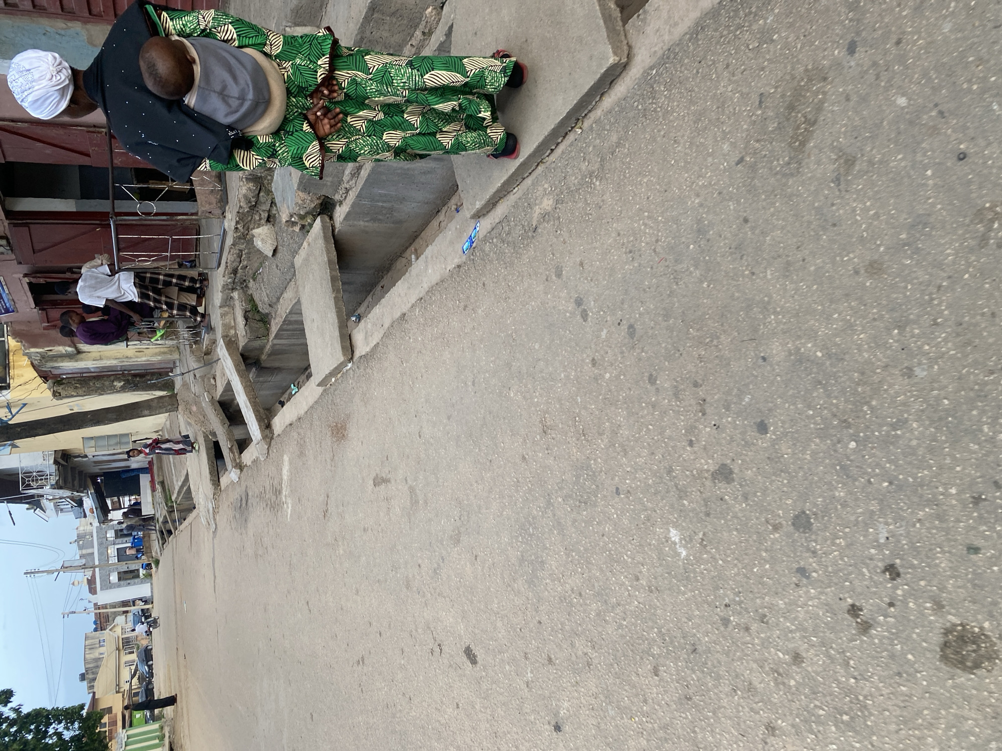 A mother wearing a green and white patterned dress and a white headscarf is seen from behind, carrying a baby in a grey carrier. She is standing on a concrete sidewalk next to a drainage ditch. In the background, a street in Jos, Nigeria, is visible, with buildings, a few cars, and people walking. The sky is overcast.