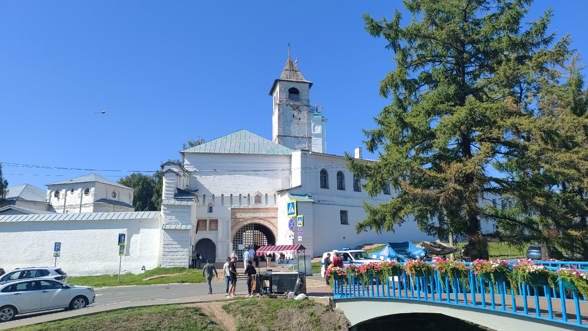 This image captures a sunny day scene in Yaroslavl, Russia, featuring the historic Spaso-Preobrazhensky Monastery. Dominating the frame is a large, white, fortified building with a prominent bell tower topped with a wooden spire. The building has a pale green roof and arched windows. In front of the building, a large arched gateway is visible, partially obscured by a canopy structure and some people. A white wall with crenellations extends to the left, encompassing the monastery grounds.
Several individuals are present near the gateway and on the adjacent sidewalk. Some appear to be walking, while others are gathered around the canopy. A police car with flashing lights is parked nearby, along with another vehicle. To the right, a bright blue bridge adorned with flower boxes filled with pink and white flowers spans a canal or river. A large evergreen tree with lush green foliage stands tall on the right side of the image, partially obscuring the monastery building. In the foreground, a road runs parallel to the monastery wall, with two cars parked on it. The sky is a clear, vibrant blue.