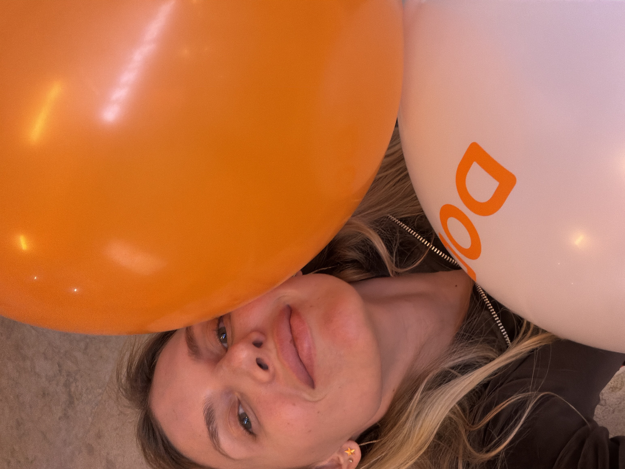 A woman lies on a carpeted floor with her head positioned between an orange balloon on the left and a white balloon with an orange "Do" logo on the right. The balloons partially obscure her face, but her eyes, nose, mouth, and chin are visible. She has blonde hair and is wearing a dark-colored jacket. Her face is tilted slightly. It is not possible to identify the location in the city of Budva, Montenegro, based on the image.