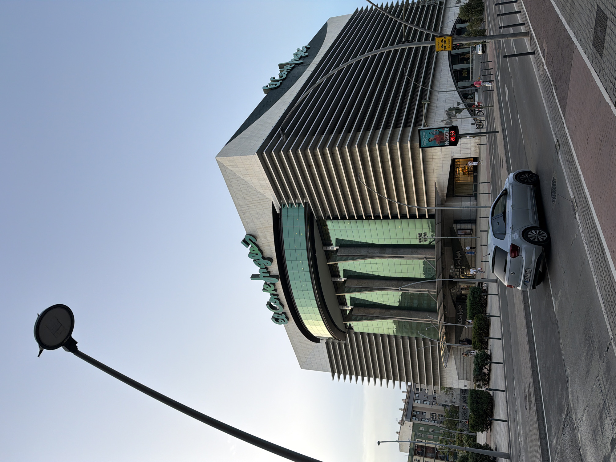 The image shows a wide street in front of a large, modern building with "El Corte Inglés" signage. The building has a distinctive architectural design with curved glass facades and horizontal louvers on its upper sections. A silver hatchback car is driving on the street in the foreground. A tall lamppost with a circular light fixture is visible on the left side of the frame, extending upwards towards a clear sky. In the background, there are other buildings, trees, and people walking on the sidewalk in front of the El Corte Inglés store. A digital billboard advertising Nescafé is mounted on the side of the building, displaying a time of "20:53". The scene appears to be in a city, possibly during the late afternoon or early evening. This is the El Corte Inglés department store located in Castelló de la Plana, Spain, on Calle de Pintor Sorolla.