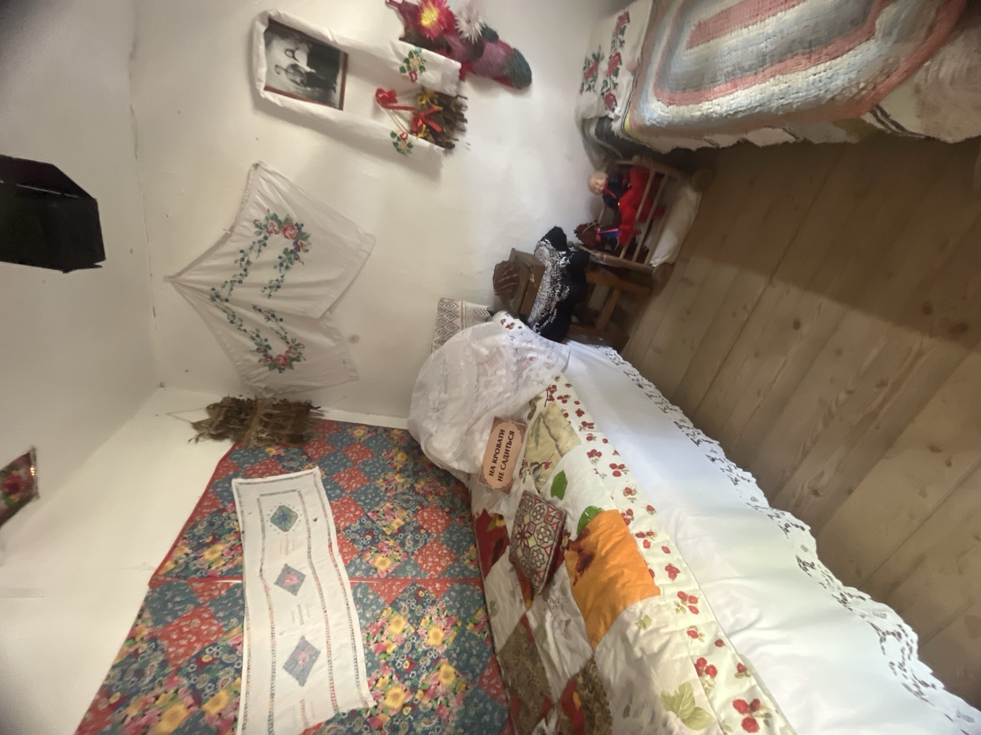 This image depicts a room decorated in a traditional, folk style, possibly within a museum or historical house in Taman', Russia. The room contains two beds, both covered with quilted blankets and decorative textiles. The bed on the left is a twin-sized bed with a white bed skirt featuring lace trim. It is adorned with a patchwork quilt in floral and geometric patterns, with a bright orange fabric swatch and a patterned cushion placed on top. A sign with Cyrillic text is visible on the bed. A draped white textile, possibly a mosquito net, is also present.
The walls are decorated with various items. The wall behind the left bed is covered with a large patchwork quilt or tapestry featuring a vibrant floral and geometric pattern. A long, white embroidered textile hangs horizontally above this, with three diamond-shaped embroidered motifs. To the right of this, a bundle of dried plants is suspended from the ceiling. Further to the right, a white fabric with embroidered floral designs is draped from the ceiling, and a framed photograph of two people is hung on the wall. Below this, several decorative items, including what appear to be woven items and a colorful sock or boot, are displayed.
A small wooden stool stands near the right bed, with a stack of boxes or containers on top, covered by a dark, patterned fabric. A small wooden crib with a doll dressed in red is placed near the floor. The right bed is a narrow twin bed with a woven, oval-shaped rug on top, featuring stripes of pink, gray, and white.
The floor is made of light-colored wooden planks. The overall impression is one of a preserved domestic interior, showcasing traditional crafts and decorative elements. The lighting suggests an indoor setting, with natural light possibly supplemented by artificial sources not directly visible.