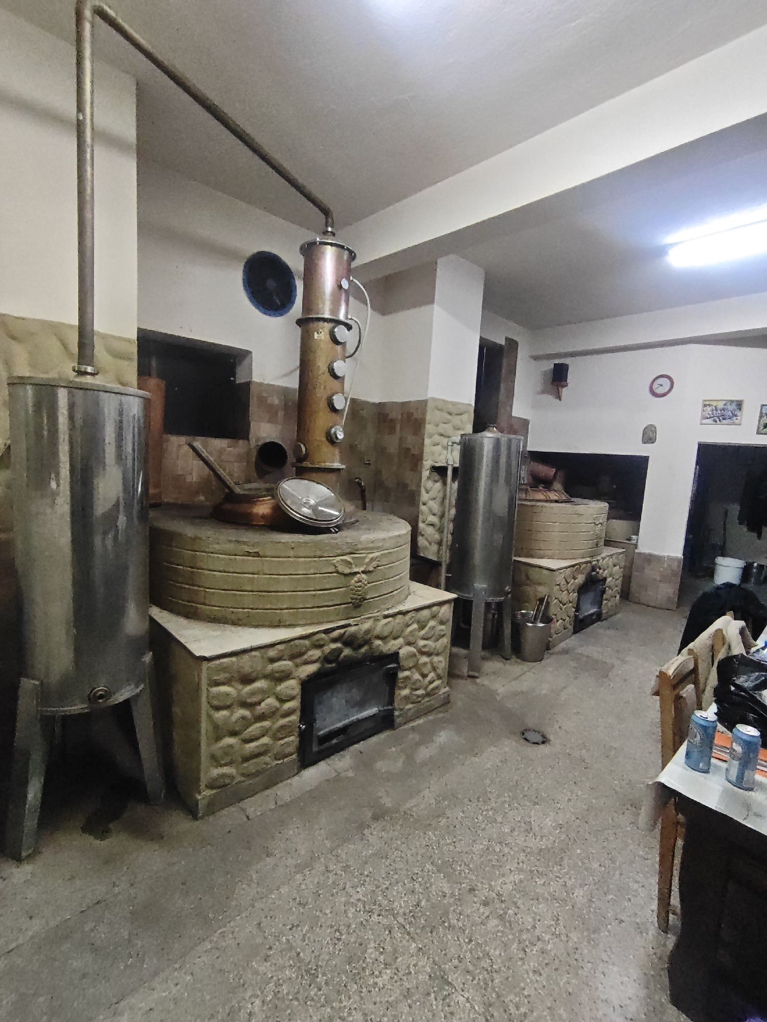 Here is a detailed description of the image:
**Scene:** The image appears to be taken inside a traditional distillery, possibly in a rural setting or small town, judging by the equipment and architecture.
**Equipment:**
* Large copper distilling apparatus dominates the scene. The apparatus features a tall pot still attached to a condenser coil, indicating it's used for distilling spirits, such as raki or brandy.
* Stone structures surround the base of the stills, likely acting as fireboxes for heating the stills.
* Large stainless steel tanks are situated nearby, potentially for storing raw ingredients or distilled products.
* Copper pot and lid sitting on the stone surface.
**Environment:**
* The room is spacious with a concrete floor.
* The walls are a mix of tiled and painted surfaces.
* The lighting is bright and appears to be from fluorescent fixtures.
* A table and chairs are present on the right side of the image, with beverage cans sitting on the table.
**Overall Impression:**
The scene suggests a functional, working distillery with a traditional setup. It does not appear to be a modern or commercial distillery but rather a more artisanal or smaller-scale operation.