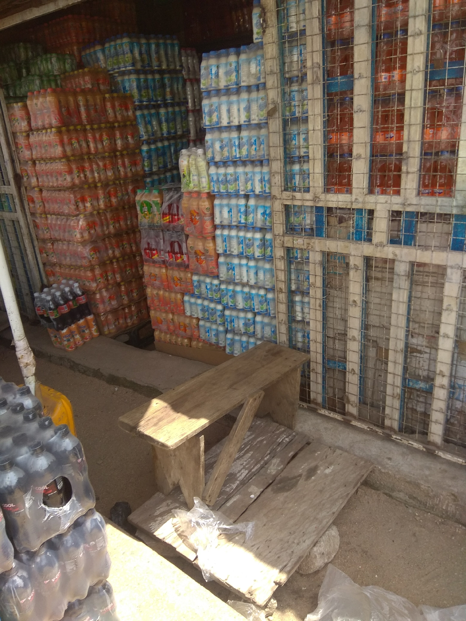 The image depicts an outdoor market stall in Jalingo, Nigeria, overflowing with bottled beverages. Stacks of plastic bottles of various sizes and colors are prominently displayed, with a particular emphasis on drinks packaged in clear plastic wrap. Some bottles are large, while others are smaller, suggesting a diverse inventory. The bottles are arranged in towering piles, creating a dense visual landscape of consumer goods.
Behind the beverages, a metal grate structure acts as a barrier or enclosure for a section of the stall. Through the grate, more bottles are visible, some appearing to be darker colored liquids in transparent bottles. A wooden bench sits in the foreground, positioned on a makeshift wooden platform. The ground beneath the stall is sandy or dusty, typical of an outdoor market environment. Scattered plastic bags and what appears to be a stone or small rock are also present on the ground.
The lighting suggests it is daytime, possibly midday, given the shadows. The overall impression is one of a busy commercial area, where goods are displayed for sale in an organized, albeit somewhat rustic, manner. The density of the product implies a high volume of sales or a well-stocked vendor. No people are visible in the image, so the activity is implied rather than directly observed. The scene is a snapshot of commerce in a Nigerian locale.