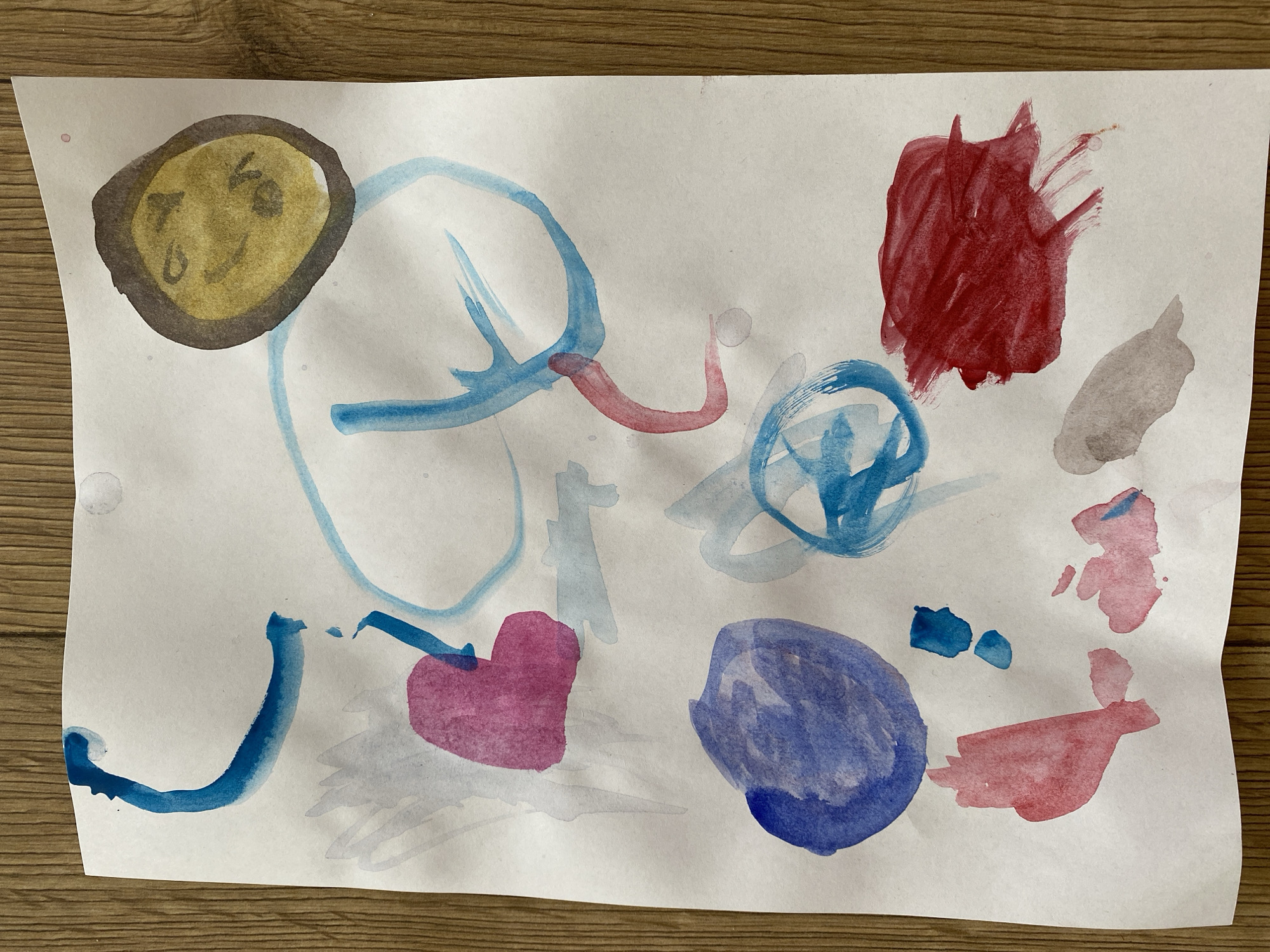 A child's watercolor painting on white paper is laid on a wooden surface. The painting features abstract splotches and lines of various colors including red, blue, pink, and yellow. Some of the shapes are round, while others are more elongated or abstract. There are no discernible figures or recognizable objects, except for a yellow oval shape with some darker markings that could be interpreted as a face. The location is unknown.