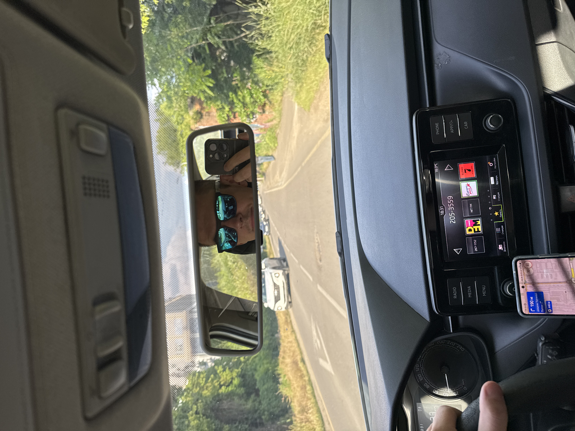 The image appears to be taken from inside a car while driving on a road lined with trees. The driver's reflection is visible in the rearview mirror. The dashboard shows a car radio display with a station playing "Europa Plus" and a navigation app on a phone mounted to the right of the radio. The navigation app shows that the car is near 190 метрах of the "улица им. Тургента", and 440 м of "Патоли". Given that "улица им. Тургента" is present, the image is in the city of Krasnodar, Russia.