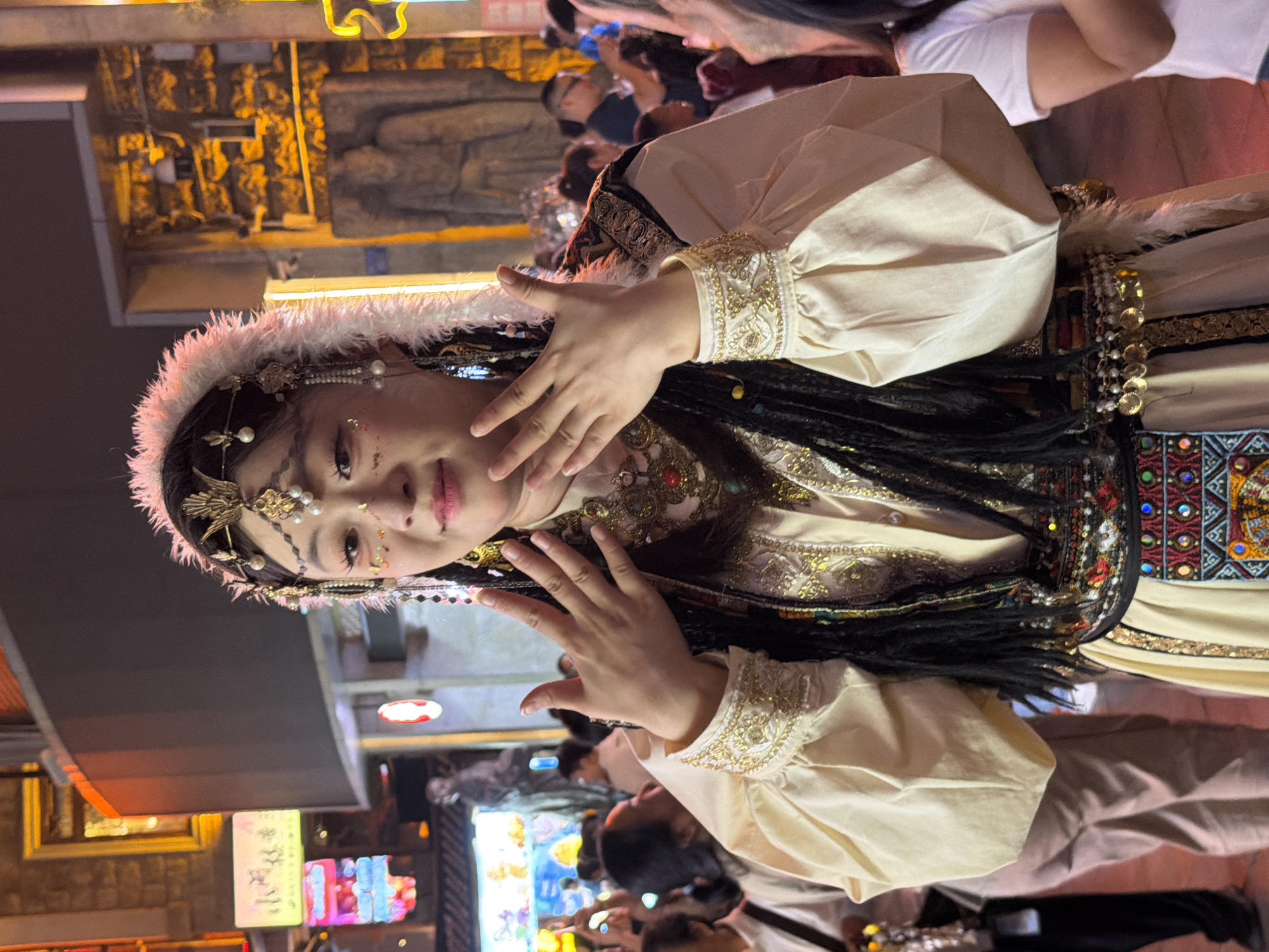In the Yuzhong District of China, a woman dressed in traditional attire stands in an outdoor setting at night. Her elaborate costume includes a cream-colored blouse with ornate gold embroidery on the sleeves and collar, a black vest with similar embellishments, and a decorative belt featuring coins and colorful patterns. She wears a feathered headband adorned with pearls and gold accents, and her hair is styled in braids. Her face is decorated with gold glitter and artistic makeup. Her hands are raised in front of her chest. The background is a bustling street scene with illuminated signs and shops, and other people are visible in the crowd, some of whom are holding up their phones. The overall atmosphere suggests a cultural event or festival.