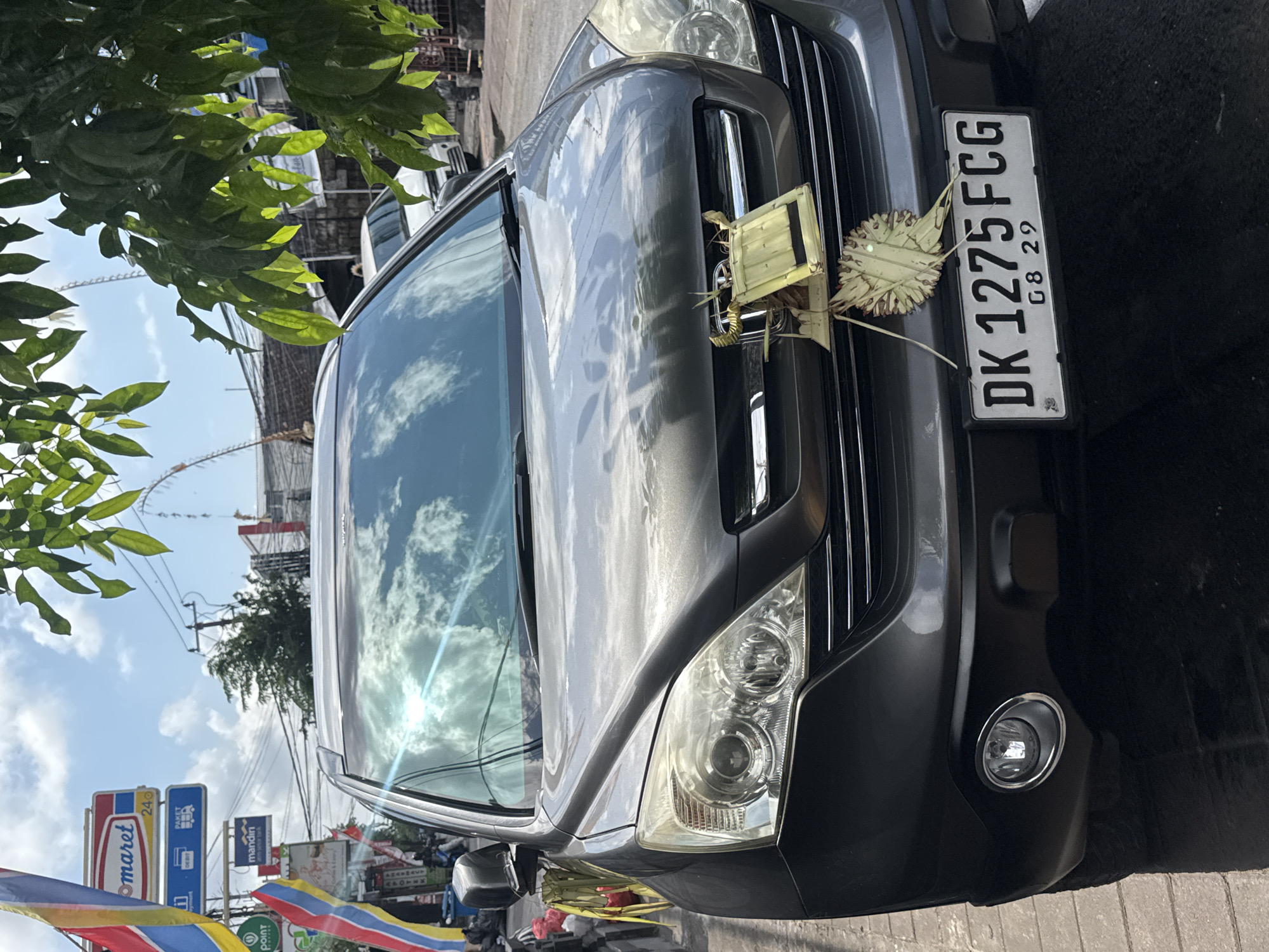 A dark grey Honda CR-V is parked on a street in Kuta, Indonesia. The car's license plate reads "DK 1275 FCG 08 29". A decorative offering made of woven palm leaves is attached to the front grille of the vehicle. The car is positioned on the right side of the street, with a sidewalk to its left. In the background, there are shops and overhead power lines. The sky is cloudy, and the sun is reflecting off the car's windshield. Red, yellow, and blue flags are visible on the left side of the image, near a sign that says "Indomaret".