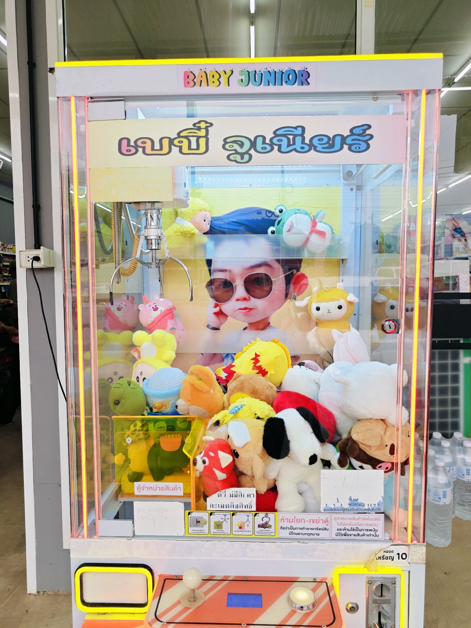 This image shows a claw machine located in Tambon Mu Si, Thailand. The machine is filled with various plush toys, including stuffed animals and cartoon characters. The top of the machine features a banner with the text "BABY JUNIOR" in English and Thai script. The machine has a clear glass front, allowing visibility of the prizes inside. A coin slot and controls are visible at the bottom of the machine. To the right of the machine, several bottles of water are stacked. The background appears to be an indoor retail environment with fluorescent lighting.