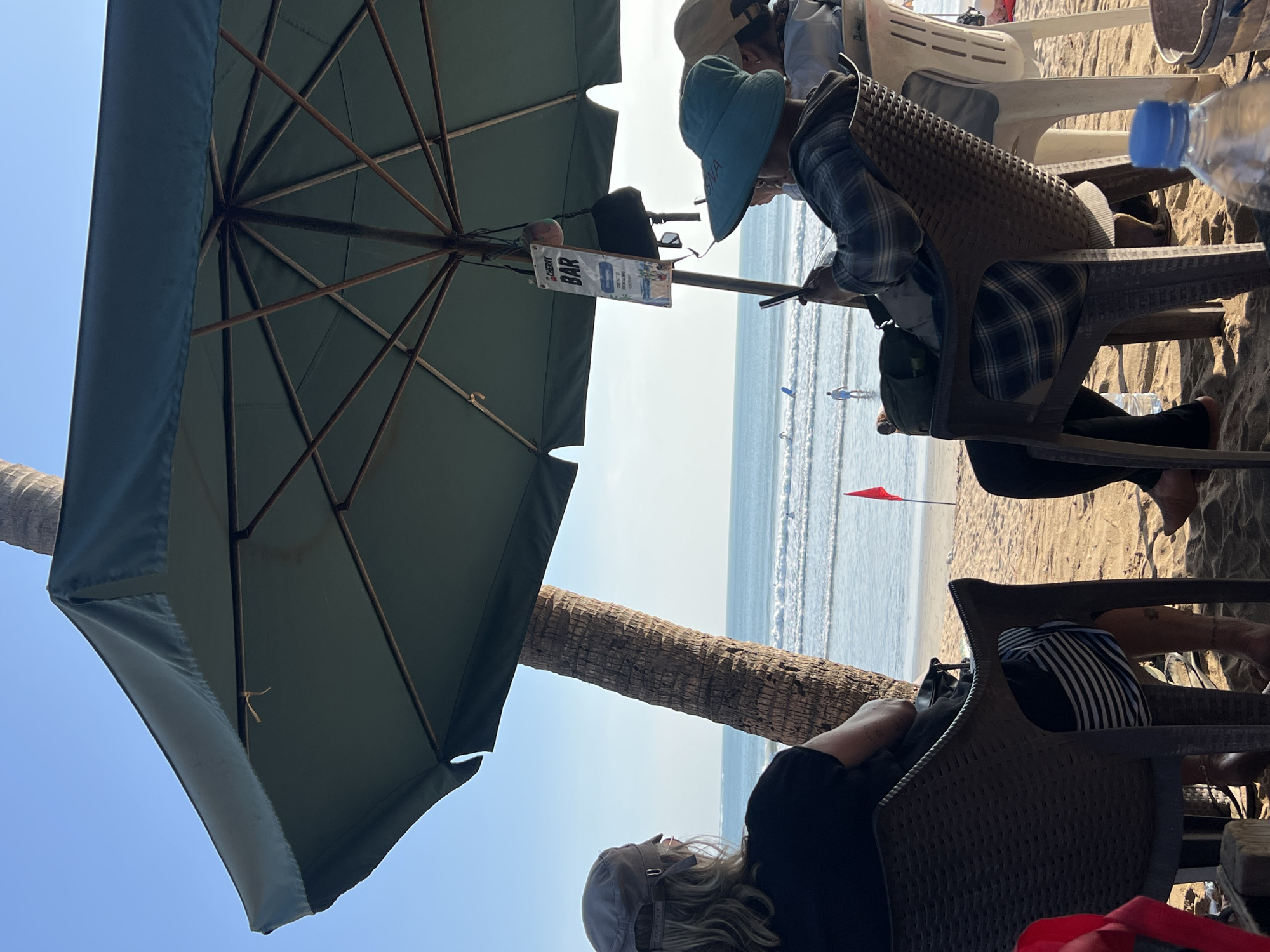 In Kuta, Indonesia, two individuals are seated under a large teal umbrella on a sandy beach, facing the ocean. A palm tree trunk is visible behind the person on the left. The ocean has gentle waves and a few distant figures can be seen in the water. A small red flag is planted in the sand near the water's edge. A sign reading "CHERRY BAR" hangs from the umbrella pole. The person on the left is wearing a cap and a black shirt, while the person on the right is wearing a blue hat and a plaid shirt. Both are looking towards the ocean. A water bottle and a bucket are visible on the sand in the foreground.