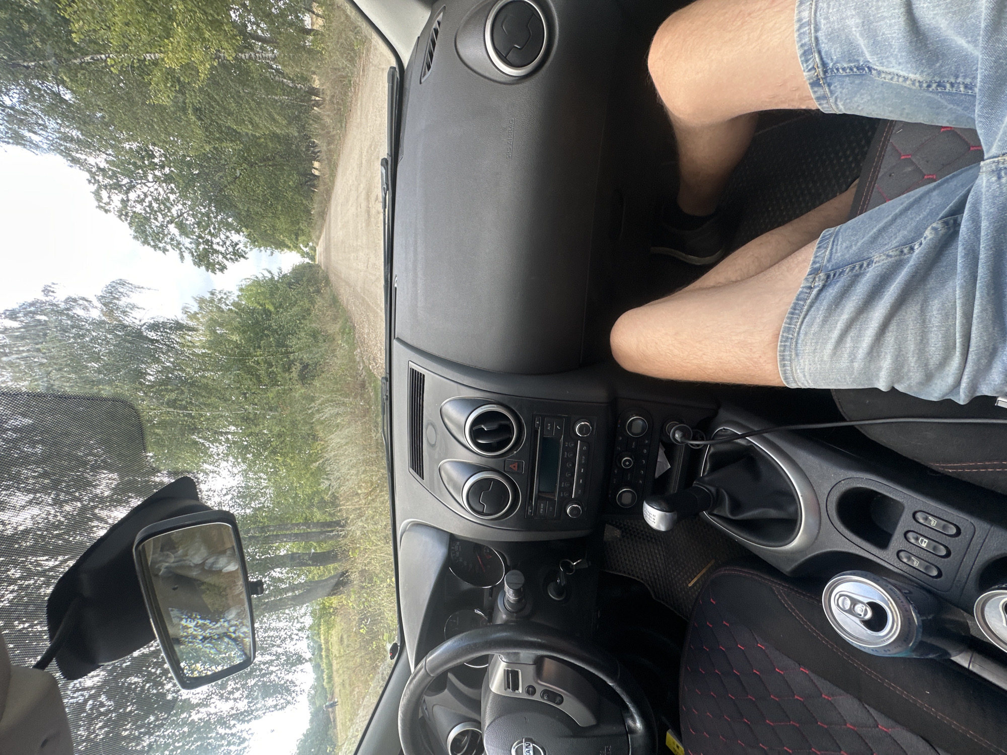 The image is a first-person perspective from inside a car driving down a dirt road lined with trees. The dashboard, steering wheel, and gear shifter are visible. A person's legs, wearing denim shorts, are in the foreground. In the background, the dirt road continues into a natural landscape with trees and foliage. A can of soda is visible in a cup holder. The car appears to be moving. The scene is outdoors on a daytime.