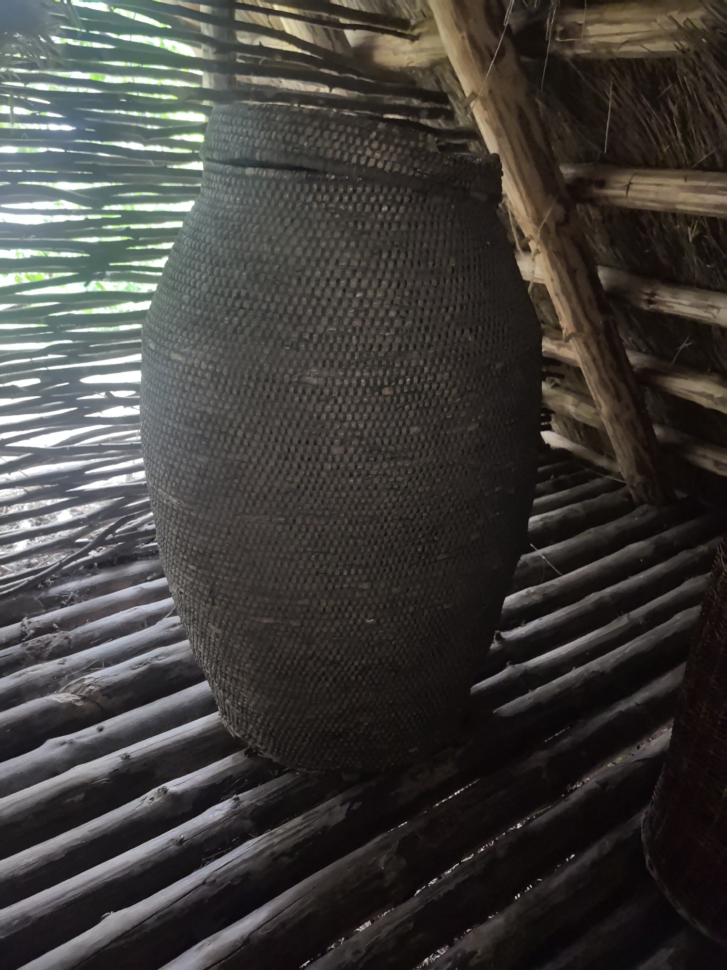 An extreme close-up shot, captured from a low angle, shows a large, dark, woven basket with a lid. The basket is positioned in the center of the frame, taking up the majority of the space. Its surface is intricately woven with a tight, repeating pattern. The basket sits on a floor constructed of horizontally laid, dark, rough-hewn logs. Above and behind the basket, the interior of a rustic structure is visible. Thin, horizontal wooden poles, likely part of a wall or screen, are visible to the left of the basket, with a soft, green, out-of-focus background suggesting foliage. To the right and above, the angled wooden beams of a thatched roof are apparent, intertwined with dried vegetation. The overall lighting is dim, with natural light filtering in from the left, highlighting the texture of the basket and the rough construction of the dwelling. The image was taken in Kamianiuki village, Brest Region, Belarus.