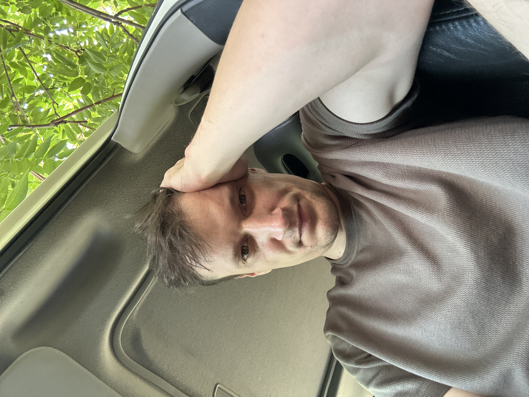 A man is shown from the chest up, looking towards the camera with his right hand resting on his forehead. He is wearing a brown t-shirt. The background shows the interior of a vehicle, with a grey headliner and a glimpse of green foliage through an open sunroof. The scene is outdoors, suggesting the vehicle is parked under a tree. The lighting is natural and appears to be daytime. There are no other people visible in the image. The location of Makiv, Ukraine, is not discernible from the visual information provided in the image.