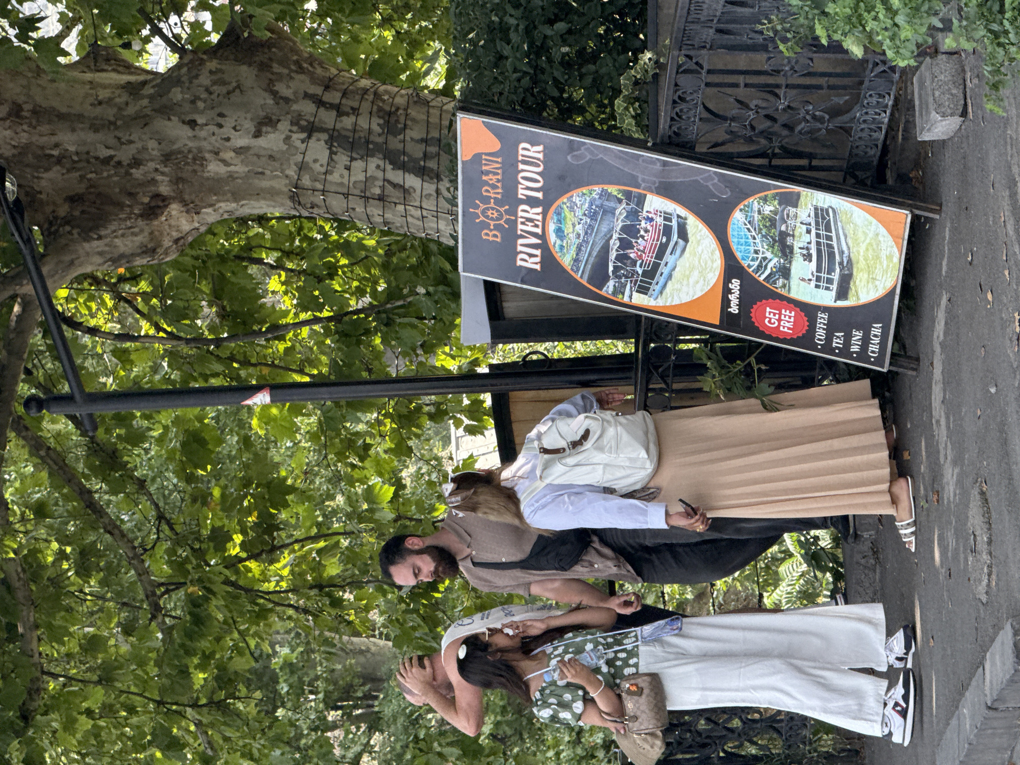 A group of four individuals, two women and two men, are standing on a paved walkway near a large tree and a black sign. The woman on the left, wearing a green and white patterned top and white pants, is holding a water bottle and appears to be talking to the man next to her. The man beside her is wearing a brown shirt and a black fanny pack. The second woman, wearing a light blue shirt and a tan pleated skirt, has a white backpack and is looking down. The fourth person, a man in the background, is wearing a white tank top and has his hand on his head. The sign, which is black with an orange border and illustrations, advertises "B-RANI RIVER TOUR" and lists "COFFEE, TEA, WINE, CHACHA" with a "GET FREE" sticker. The background shows lush green foliage and a textured tree trunk wrapped with wire. The scene appears to be outdoors, possibly in a park or along a waterfront in Tbilisi, Georgia, given the Georgian script on the sign.