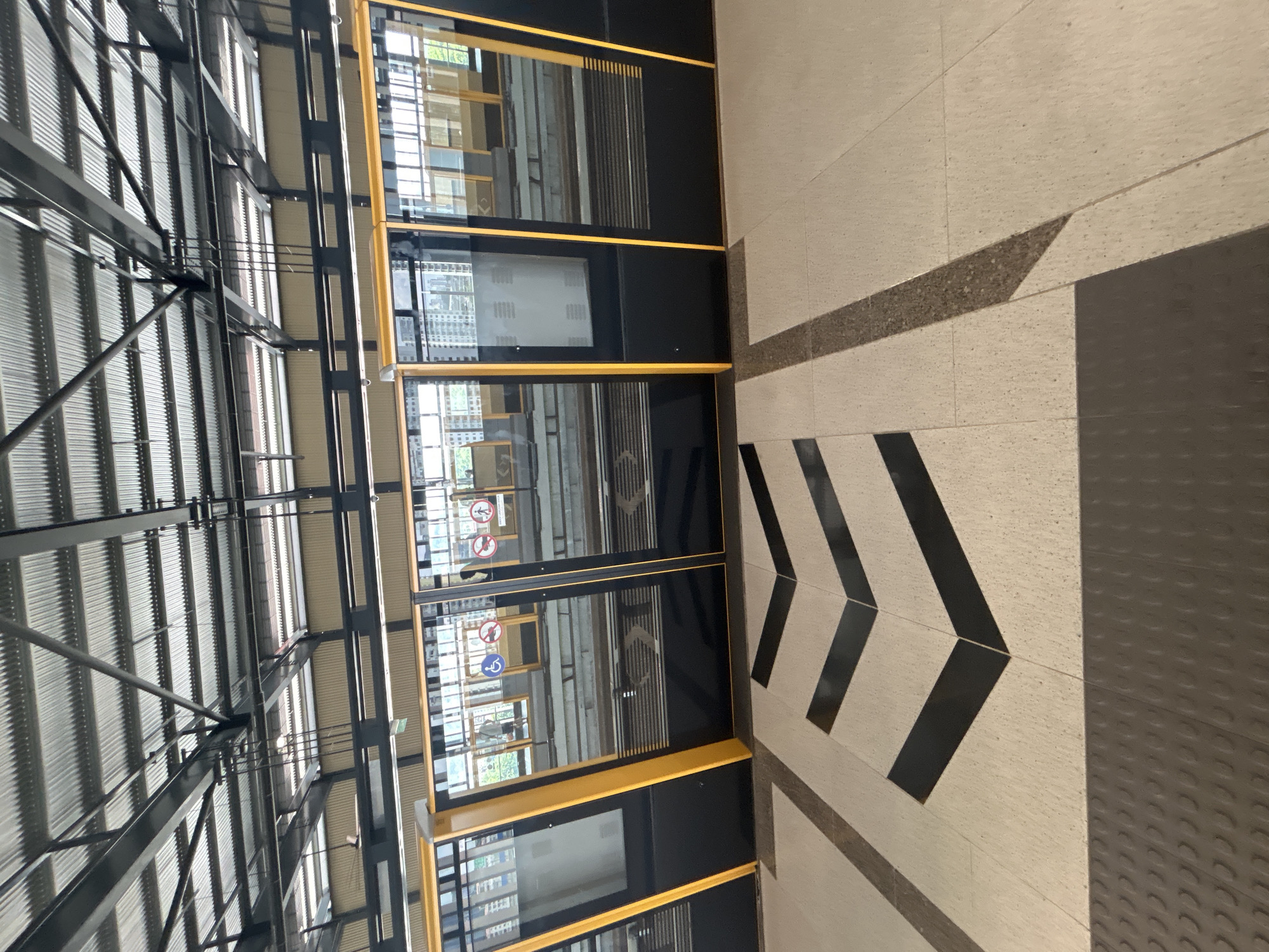 This image depicts a train platform at an urban transit station in Kuala Lumpur, Malaysia. The platform is modern, featuring a corrugated metal roof supported by a dark metal framework. Yellow-trimmed, black glass doors of a train are visible, reflecting the surrounding environment. The floor of the platform is tiled in a light color with dark, chevron-patterned markings. A tactile paving strip, designed for visually impaired individuals, runs along the edge of the platform. The train doors show various safety signage, including symbols for wheelchair accessibility and prohibitions against smoking and eating. The reflections in the doors suggest a cityscape with buildings and possibly other platforms or waiting areas. The scene is clean and well-maintained, indicative of a functional public transportation hub. There are no people clearly visible in the immediate foreground.