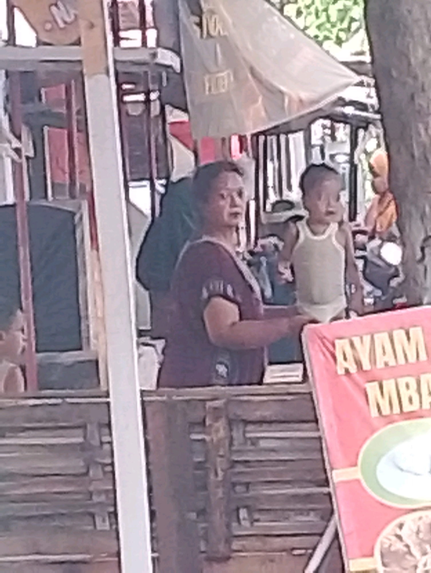 This is a medium-angle, blurry, outdoor image of a street scene, likely in Probolinggo, Indonesia. The focus is on a food stall with a pink banner displaying text in yellow that reads "AYAM MBA." In the foreground, a person is partially visible on the left, and to the right, a woman wearing a purple shirt is standing behind a wooden counter, appearing to interact with someone or something outside the frame. A young child in a light-colored tank top and underwear stands to the woman's right. Behind them, there are stalls and trees. The lighting suggests it is daytime.