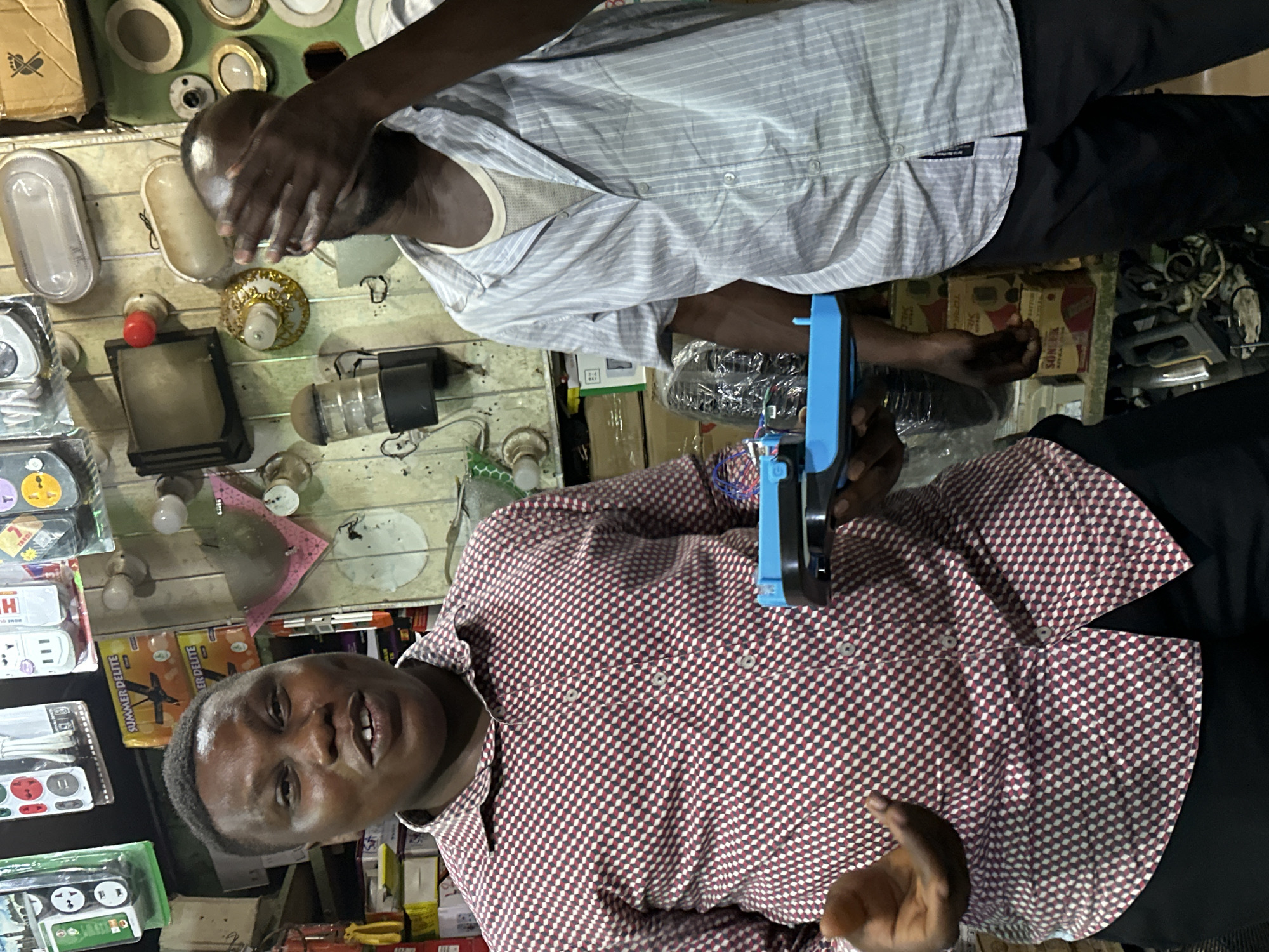 Two Nigerian men stand in what appears to be an electrical supply shop in Narayi, Nigeria. The man on the left, wearing a red and white checkered shirt, faces forward, smiling and holding a blue electronic device. The man on the right, dressed in a light blue and white striped shirt, covers his face with his hand and looks away. The shop's walls are covered with various electrical items, including light fixtures, power outlets, and boxes of supplies, suggesting a busy retail environment. The overall scene depicts a casual interaction within a marketplace setting.