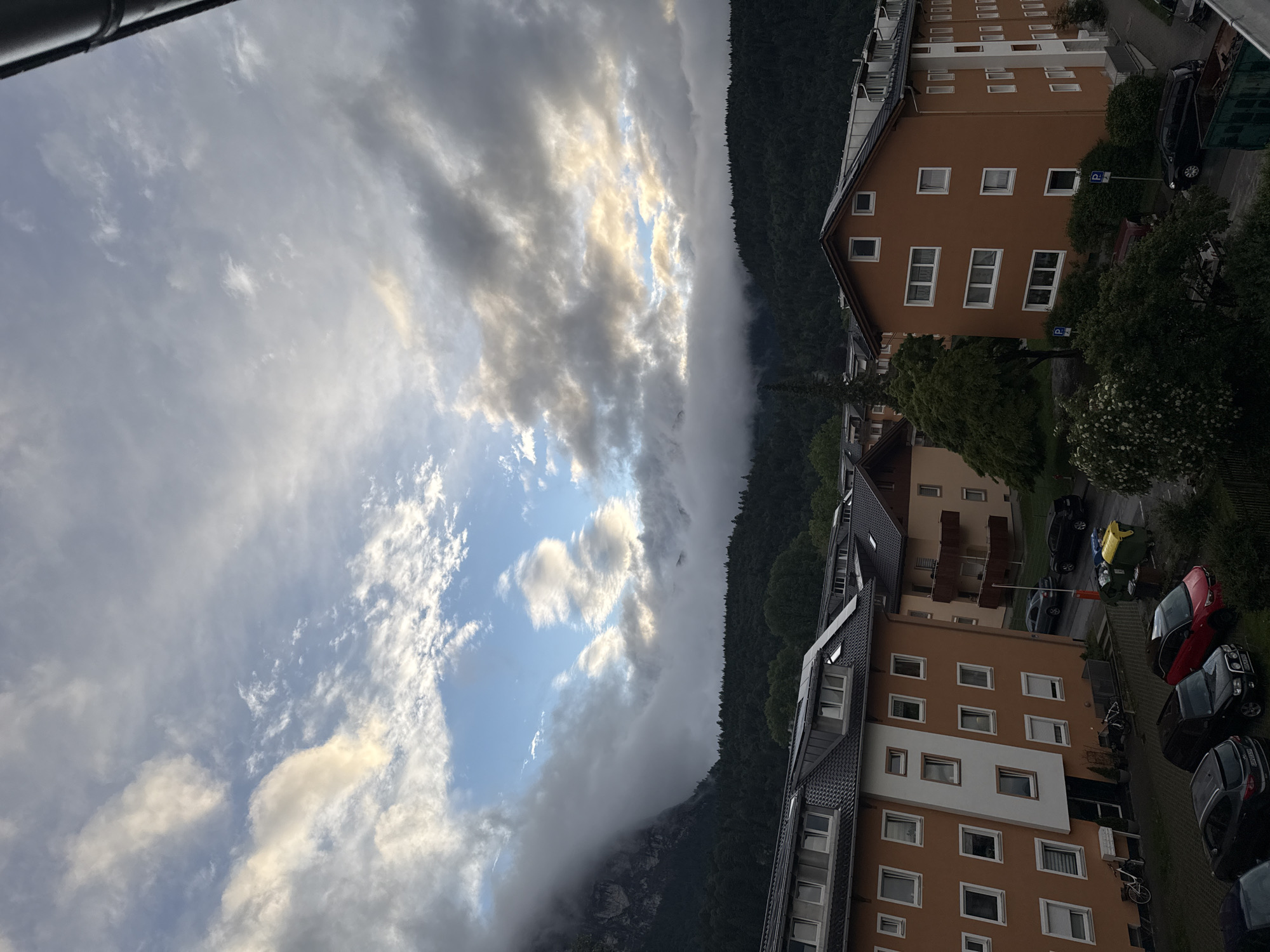 The image depicts a view of apartment buildings and a forested mountainside under a dramatic sky. The apartment buildings are modern, with orange and white facades and dark tiled roofs. Some have balconies and visible windows. In the foreground, cars are parked on asphalt and gravel surfaces. A row of trash bins is visible to the right. A dense forest covers the lower slopes of a mountain range, with some of the peaks obscured by low-lying clouds and fog. The sky is filled with a mix of light and dark clouds, with patches of blue sky and sunlight breaking through.
Based on the architectural style of the buildings, the prevalence of forested mountains, and the general landscape, this image is likely located in Garmisch-Partenkirchen, Germany. The scene appears to be taken during late afternoon or early evening, given the lighting conditions and cloud cover. There are no people visible in the image. The overall impression is one of a somewhat overcast day in a mountainous town.