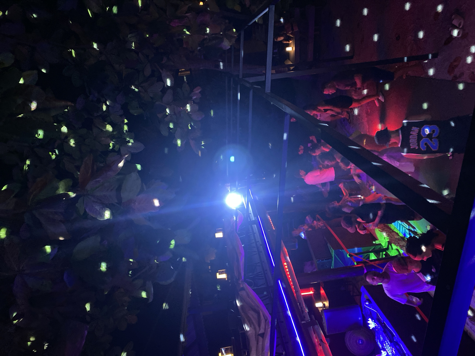 A low-angle shot captures a party scene in Ban Haad Rin, Thailand. The foreground displays lush foliage with sporadic bright lights, suggesting decorative lighting. In the background, a group of people is gathered on a raised platform. One individual wears a "Jordan 23" jersey. A structure with blue and red neon lights bisects the scene, adding to the vibrant nightlife atmosphere. Above, disco ball reflections speckle the ceiling.