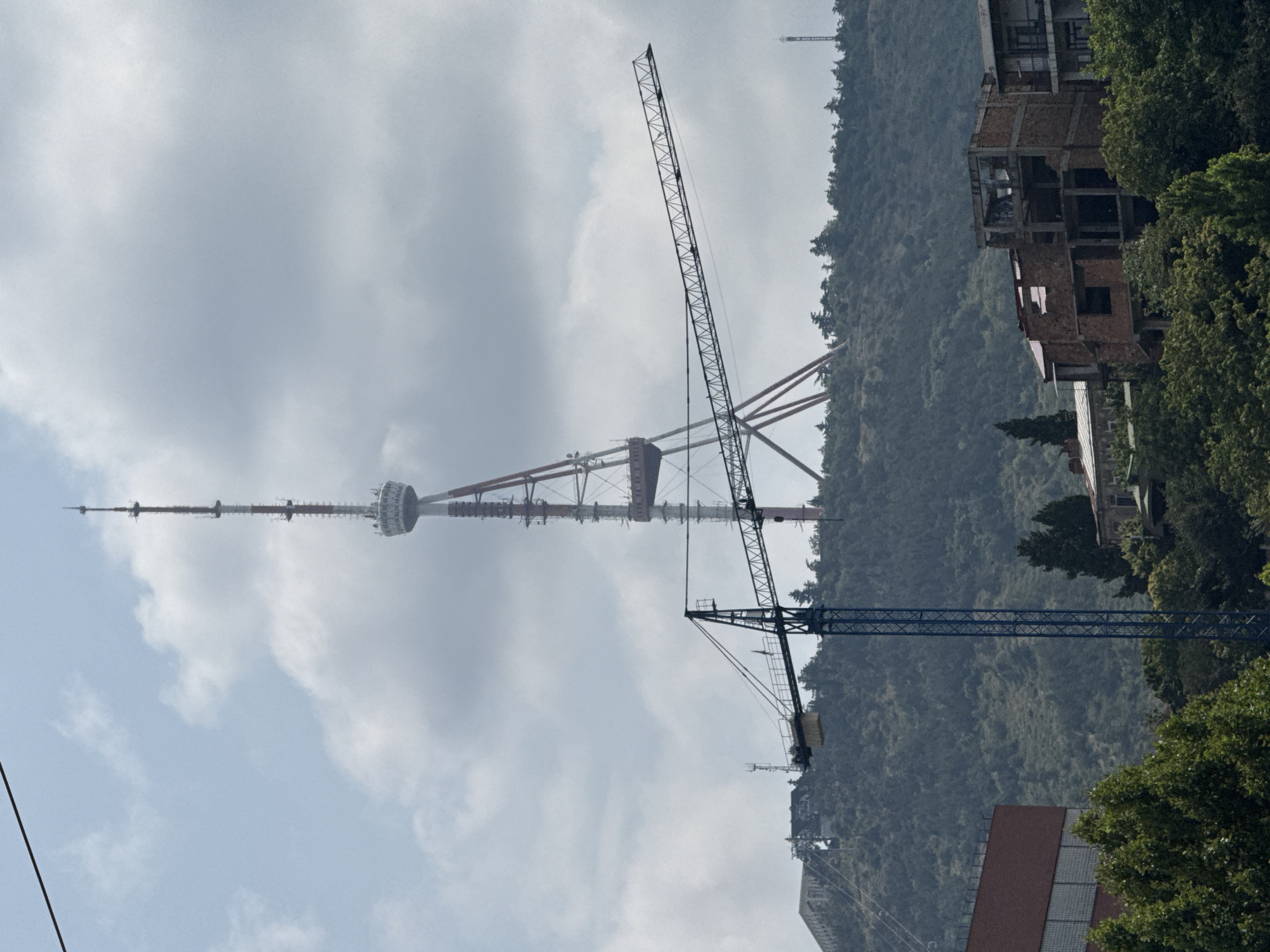 The image captures a distant view of the Tbilisi TV Tower, a prominent landmark situated on Mount Mtatsminda in Tbilisi, Georgia. The tower, a tall, lattice structure with a white and red design, stands prominently against a cloudy sky. In the foreground, a construction crane, predominantly blue, dominates the mid-ground. Its long jib extends horizontally across the frame, positioned in front of the TV tower. The base of the crane is visible, with its vertical structure extending upwards.
Below the crane and to the right, several buildings are visible, some appearing to be under construction, with exposed brickwork and incomplete structures. To the left, a red building with reflective windows is partially visible. The background consists of a densely forested hillside, indicating the natural landscape surrounding the city. The overall scene suggests a blend of urban development and natural scenery, with a focus on the construction activity and the iconic TV tower. The lighting suggests it is daytime, with diffused sunlight due to the overcast conditions. There are no people visible in the image.