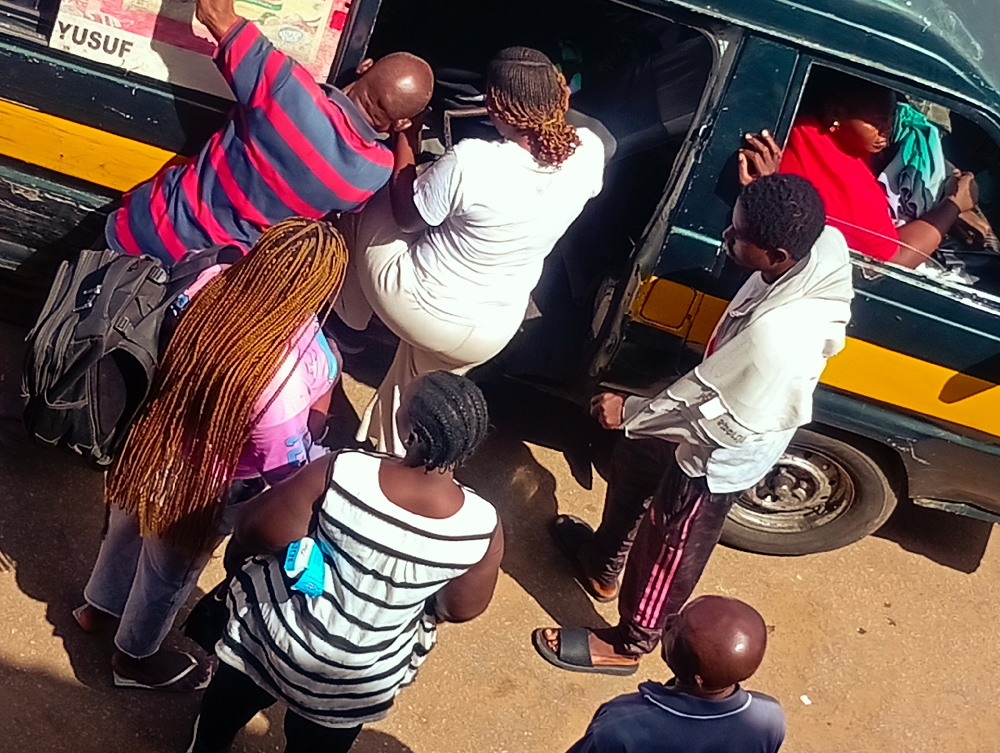 In Bukuru, Nigeria, a group of people are gathered around the open door of a dark green van. A man wearing a red and blue striped shirt is leaning into the van, his arm extended. Beside him, a woman with long, braided brown hair is standing, and a portion of a pink shirt is visible. A black backpack rests on the ground near them. Inside the van, a person in a bright red top is looking out the window, their hands resting on the door frame. Another person, wearing a white garment, is partially visible inside the van. In the foreground, a woman in a black and white striped top stands, facing away from the camera, and to her right, a man in a blue shirt and a dark-haired man wearing a light grey shirt and dark pants with pink stripes are visible. The ground beneath them appears to be dusty earth. A yellow stripe runs along the lower portion of the van.