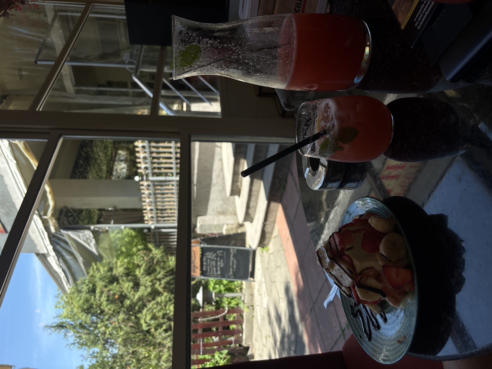An outdoor cafe table is set with a dessert and drinks, likely in Plovdiv, Bulgaria. The dessert is a mound of what appears to be ice cream or a panna cotta, topped with sliced strawberries, bananas, and drizzled with chocolate sauce. It sits on a teal-colored plate with a fork on the left side. Adjacent to the dessert is a clear glass filled with a pinkish-red beverage, adorned with mint leaves and a straw, with ice cubes visible. A tall glass carafe, also containing the pinkish-red drink, stands to the right of the glass. The table surface is dark and reflective, showing the reflections of the objects on it. A metal ashtray is positioned near the glass. Through the cafe's window or open side, a sunlit outdoor area is visible, featuring greenery, a wooden fence, stone steps, and a chalkboard sign. The architecture outside suggests a European setting, possibly Plovdiv. The lighting is natural, with sunlight casting shadows. No people are visible in the frame.