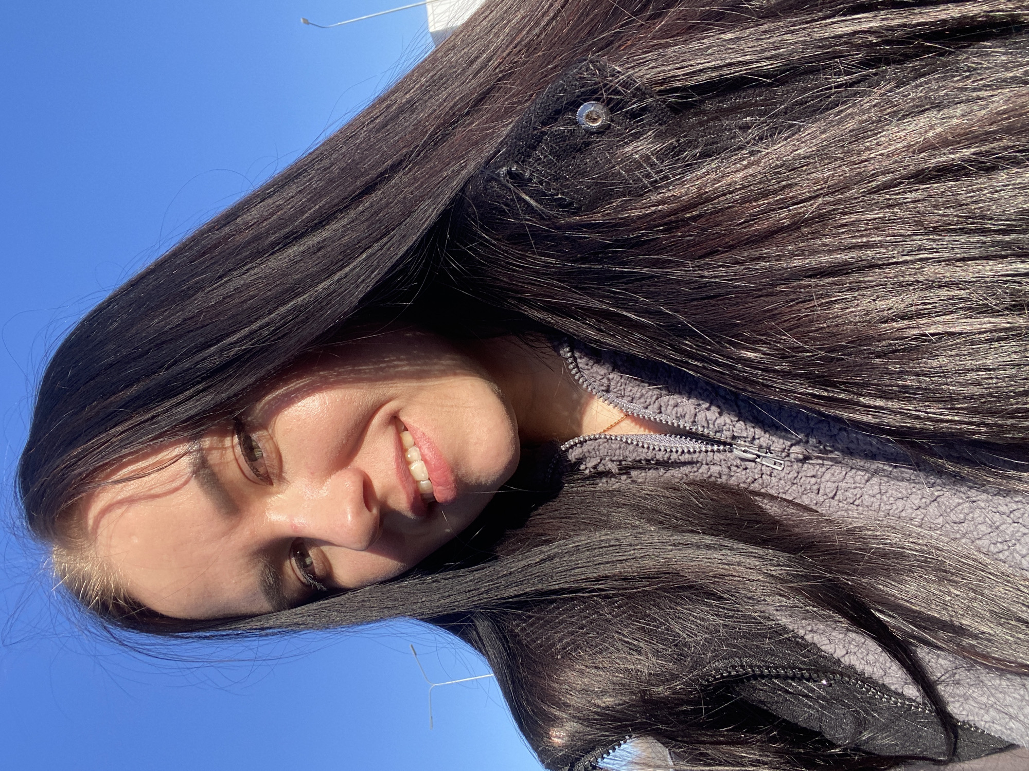 A woman with long, dark hair and light skin smiles towards the camera against a clear, blue sky. She wears a grey, fleece jacket with a zippered closure. The exact location within the city of Votkinsk, Russia, cannot be determined from the image alone.