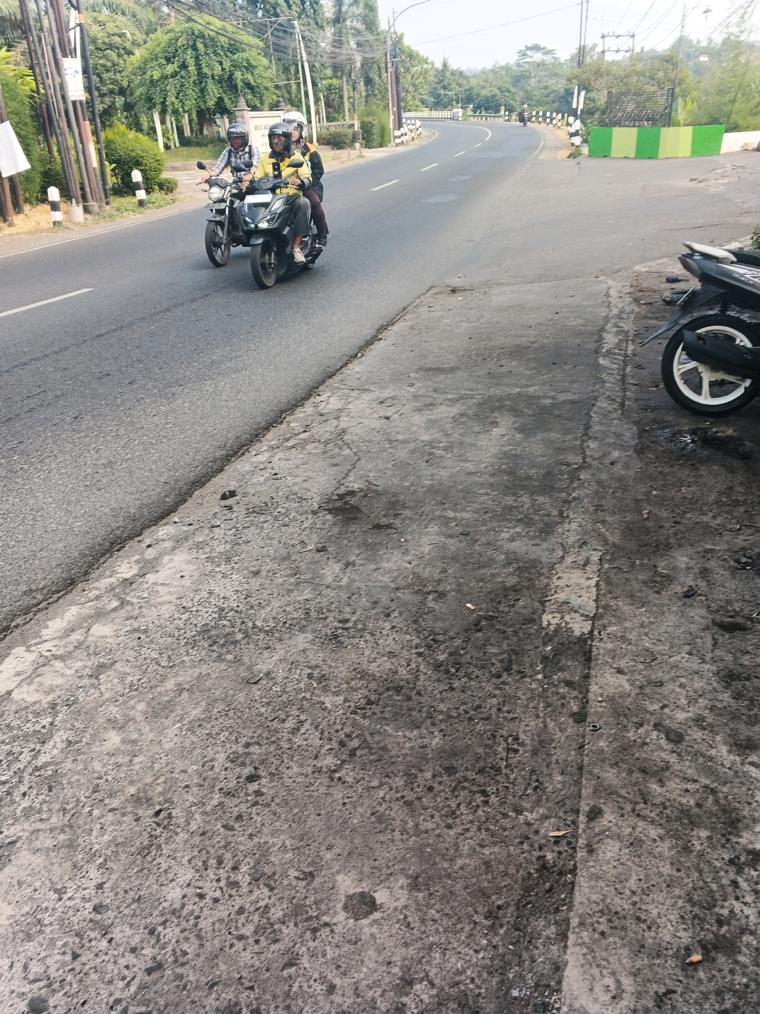 Two individuals on a motorcycle, one driving and one as a passenger, are traveling on a paved road in Ungaran, Indonesia. The driver wears a yellow shirt and a black helmet, while the passenger is dressed in black and wears a white helmet. They are proceeding down a curving road bordered by trees on the left and a low wall with green and yellow panels on the right. In the foreground, a portion of the roadside is visible, composed of rough, gray concrete with some patches of dirt and debris. To the far right, the rear wheel of a parked motorcycle is visible. The sky is bright, indicating daytime.