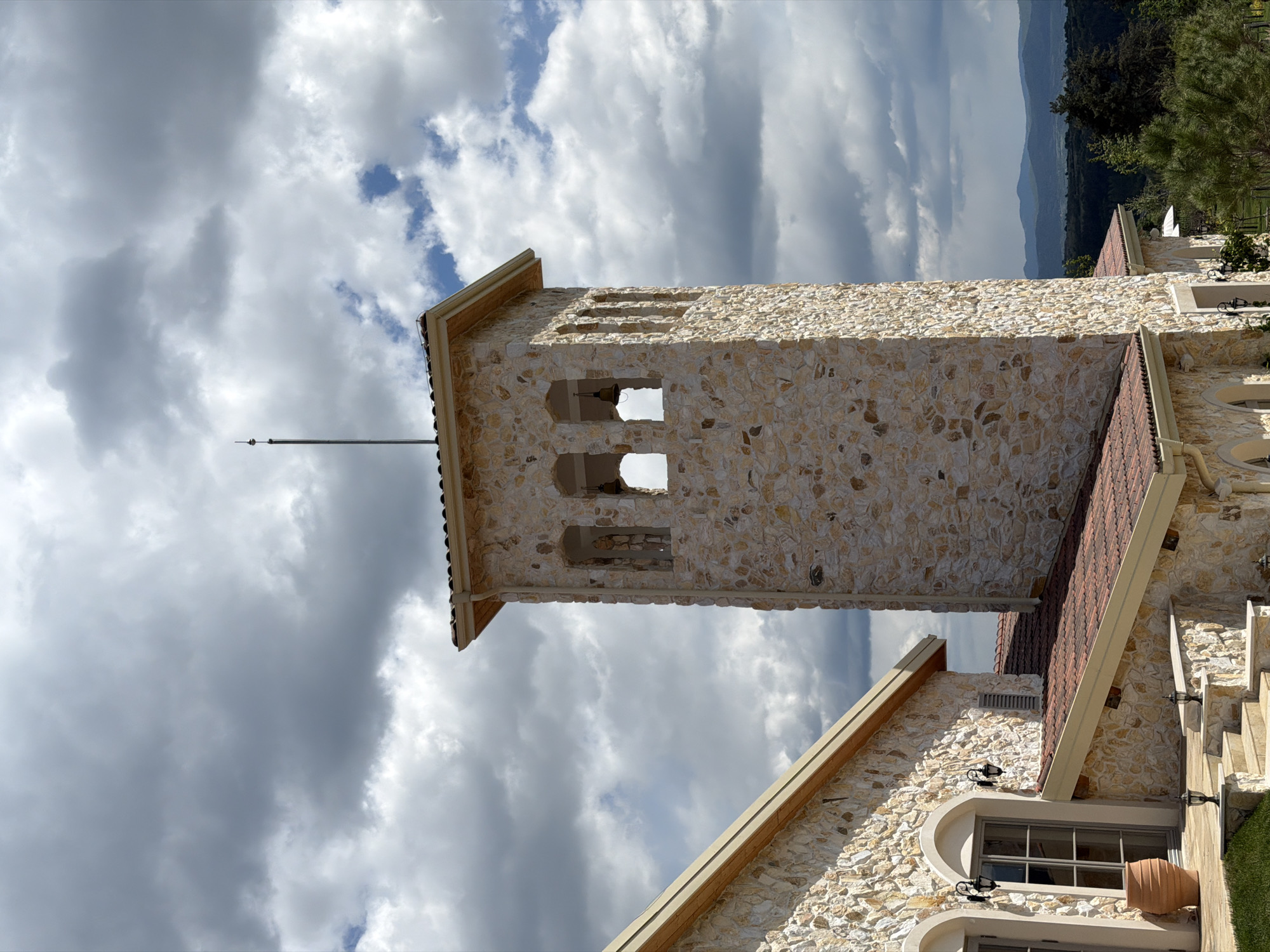The image shows a stone tower with a metal lightning rod attached to the top. The tower has three open archways on its upper portion. The tower is part of a stone building with a red tile roof. The building appears to be located in Kocherinovo, Bulgaria. The sky is cloudy.