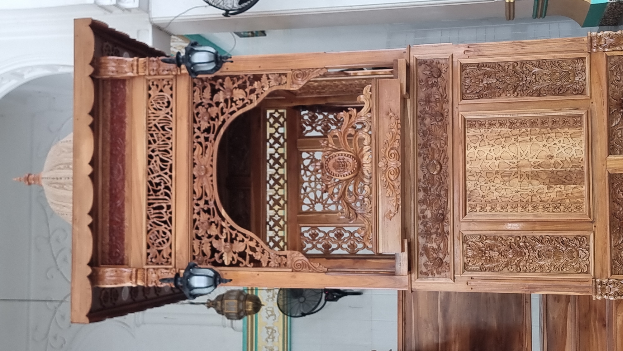 The image shows a close-up, frontal view of a highly ornate, wooden minbar, or pulpit, situated in a mosque in Lapang Timu, Indonesia. The minbar is constructed from dark wood and features intricate carvings throughout. At the top, a decorative canopy is supported by carved pillars. The central section of the minbar is arched and features geometric patterns and Arabic calligraphy carved into the wood. Below this, a decorative panel displays a circular motif with floral elements. The lower part of the minbar is divided into panels, with some featuring floral carvings and others displaying geometric patterns. Two decorative lanterns are suspended from the sides of the upper structure. In the background, a portion of the mosque's interior is visible, including white walls with decorative molding and a green stripe along the top. A fan is also visible on the left side of the image. The lighting is bright, suggesting an indoor setting during the day.