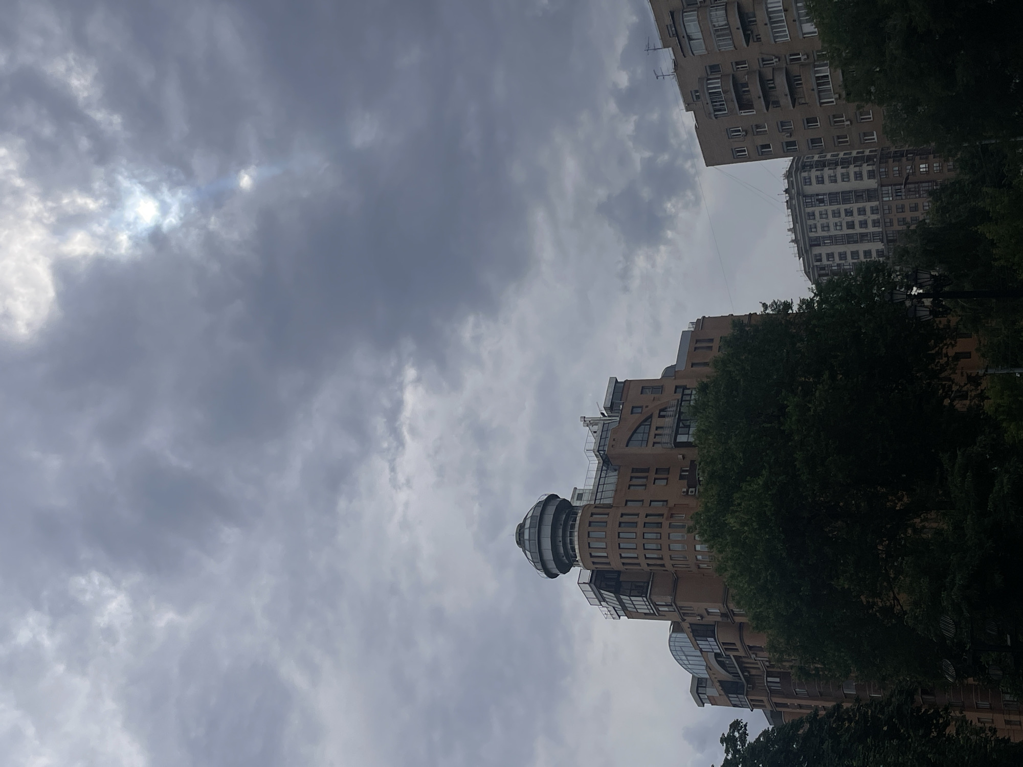 This is a low-angle, outdoor, eye-level shot of a cityscape in Moscow, Russia. The scene is dominated by overcast skies with patches of sunlight breaking through, suggesting it is daytime. The weather appears cloudy or possibly during a light shower.
The primary subjects are several modern residential buildings. One building on the right features a distinctive, rounded, glass-enclosed structure on its top. The buildings are made of light-colored brick and have numerous windows, balconies, and architectural details. Lush green trees are interspersed among the buildings, obscuring some of the lower portions of the structures and the ground.
The image conveys a sense of urban living within a somewhat subdued, cloudy atmosphere. There are no people visible in the frame. The overall mood is neutral, focusing on the architectural elements and the sky. The location is identifiable as Moscow, Russia, due to the architectural style of the buildings and the context provided. No text is visible in the image.