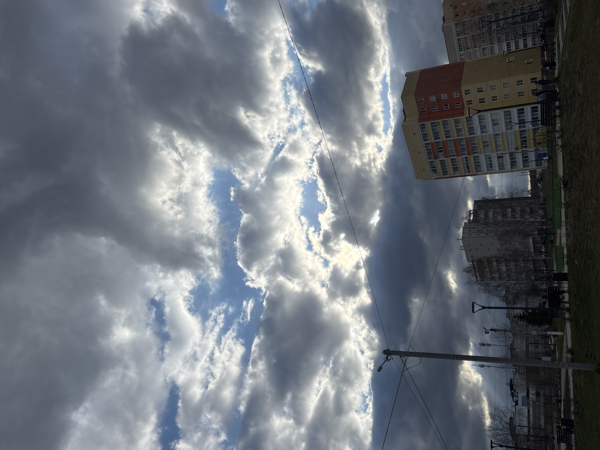 This image captures a dynamic urban sky and cityscape in Chaikovsky, Russia. The dominant feature is a dramatic sky, filled with large, dark grey clouds that stretch across the frame. Bright sunlight breaks through these clouds in several places, creating brilliant white patches and revealing glimpses of blue sky, suggesting either the sun is behind the clouds or it's a day with broken cloud cover.
On the right side of the frame, several multi-story residential buildings are visible. The most prominent is a colorful building with sections painted in yellow, orange-red, and grey/white, featuring numerous balconies. Above it, another, older grey building is partially visible. The ground below these buildings appears to be a strip of grassy, somewhat unkempt area, indicative of a park or courtyard. Bare trees, possibly signifying late autumn or early spring, are scattered across the scene.
A tall concrete utility pole with power lines extends from the bottom right into the mid-frame, adding an industrial element to the urban landscape. Several streetlights and park benches are also present. Small figures of people can be observed near the colorful building, suggesting typical daily activity.
Visible text includes a yellow pedestrian crossing sign and another small yellow sign, possibly indicating parking, on the facade of the colorful building. The time of day appears to be daytime, with the dramatic lighting suggesting either late morning or late afternoon. The overall atmosphere is one of an active urban environment under a powerfully expressive sky.