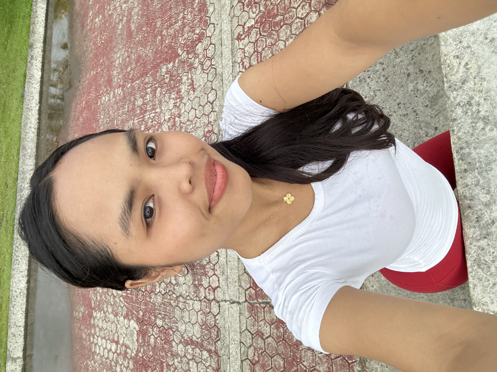 A selfie-style portrait features a young woman with a pleasant, subtle smile, her lips lightly parted to reveal pink lipstick. She has dark hair, styled down, and dark eyes. She is wearing a simple white short-sleeved t-shirt, a gold-colored necklace with a four-leaf clover pendant, and small hoop earrings. A glimpse of a red garment, possibly a top or a skirt, is visible beneath her white shirt.
The background suggests an outdoor setting, possibly a park or public area in Davao City, Philippines. The ground surface features a distinctive pattern: a reddish-brown, textured area resembling a honeycomb or hexagonal grid, which appears somewhat worn or faded in parts. To the left, a strip of green grass or foliage is visible, alongside a grey concrete border that looks slightly wet. Another section of grey, speckled concrete is seen towards the right. The lighting suggests it is daytime. The woman's expression conveys a calm and amiable mood. No specific activity is depicted other than posing for the photo. There is no visible text in the image.
