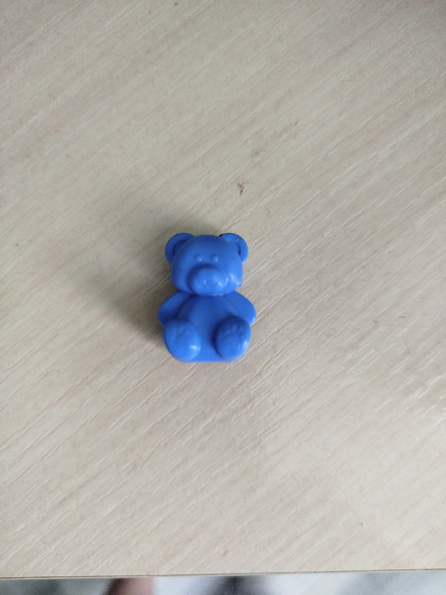 A small, blue, plastic teddy bear figurine is centered on a light-colored, wood-grained surface. The surface has diagonal lines, suggesting a wooden table or countertop. The lighting creates a soft shadow to the left of the bear. The scene is indoors and appears to be a still life. There is no indication of the specific location within Крывасельскі сельскі Савет, Belarus, as the image only depicts the object and its immediate surroundings.