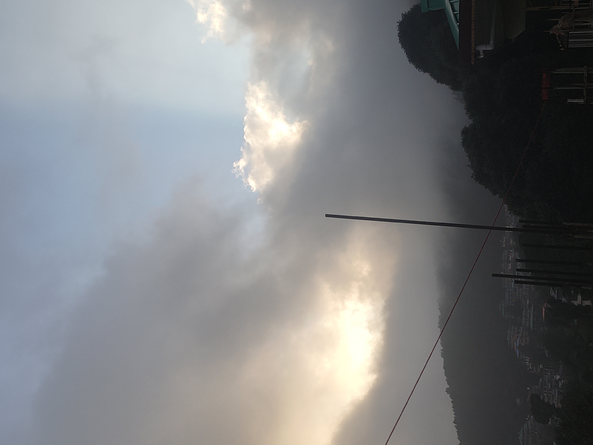A low-angle, vertical shot captures a cloudy sky over a misty mountainous landscape. The sky is a gradient of light blue at the top, transitioning to lighter shades of gray and white as it moves down. Sunlight breaks through the clouds in the upper right quadrant, creating a bright, luminous patch. A thin, dark pole extends vertically from the bottom center of the frame upwards, partially obscuring the view of the sky. In the distance, a town or village is nestled on the side of a hill, with buildings appearing as small, colorful specks due to the mist and distance. The hillside is covered in dark, dense trees. A red wire is strung diagonally across the lower left portion of the image. The overall mood is atmospheric and slightly mysterious, due to the prevalent mist and overcast sky. The location is identified as Dharamkot, India.