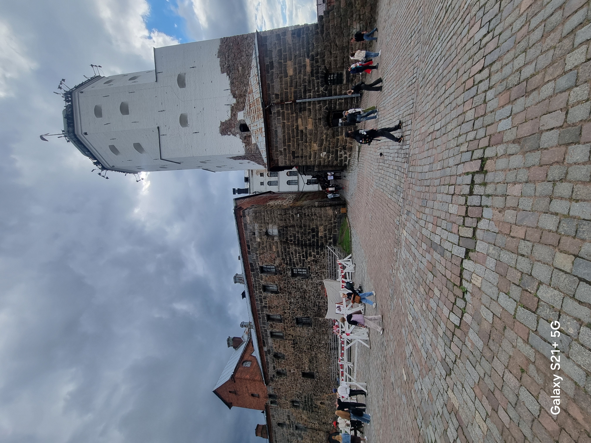 The image shows Vyborg Castle in Vyborg, Russia, under a cloudy sky. The castle features a tall, white tower with a dark, stone base. To the left of the tower is a red-brick building with a gray roof, followed by a long, dark stone building with numerous windows. The ground in front of the castle is paved with cobblestones. Several people are scattered across the plaza, some walking, others standing in small groups. In the foreground, a person with a backpack walks away from the camera. There are white barricades with red and white flags set up in front of the stone building. The overall scene depicts a historic landmark on a somewhat overcast day, with a few visitors present.