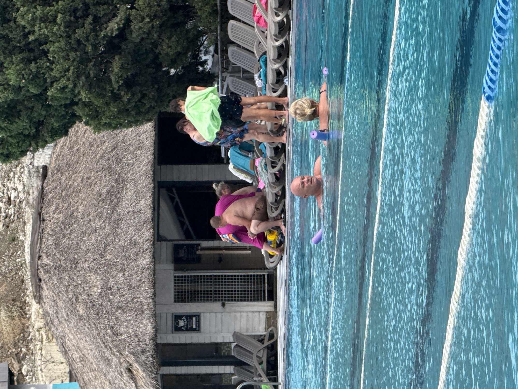 In the foreground, a man and a woman are partially submerged in a swimming pool, both holding purple flotation devices. The pool water is a bright turquoise and divided into lanes by white ropes. In the background, a thatched-roof building with an open entrance is situated behind a paved area with several lounge chairs. A group of people are gathered near the building, some appearing to be drying off with towels. Behind the building, a large, lush green tree stands tall. The overall scene suggests a recreational setting, likely a resort or public pool, in Массандровский поселковый совет, Ukraine.