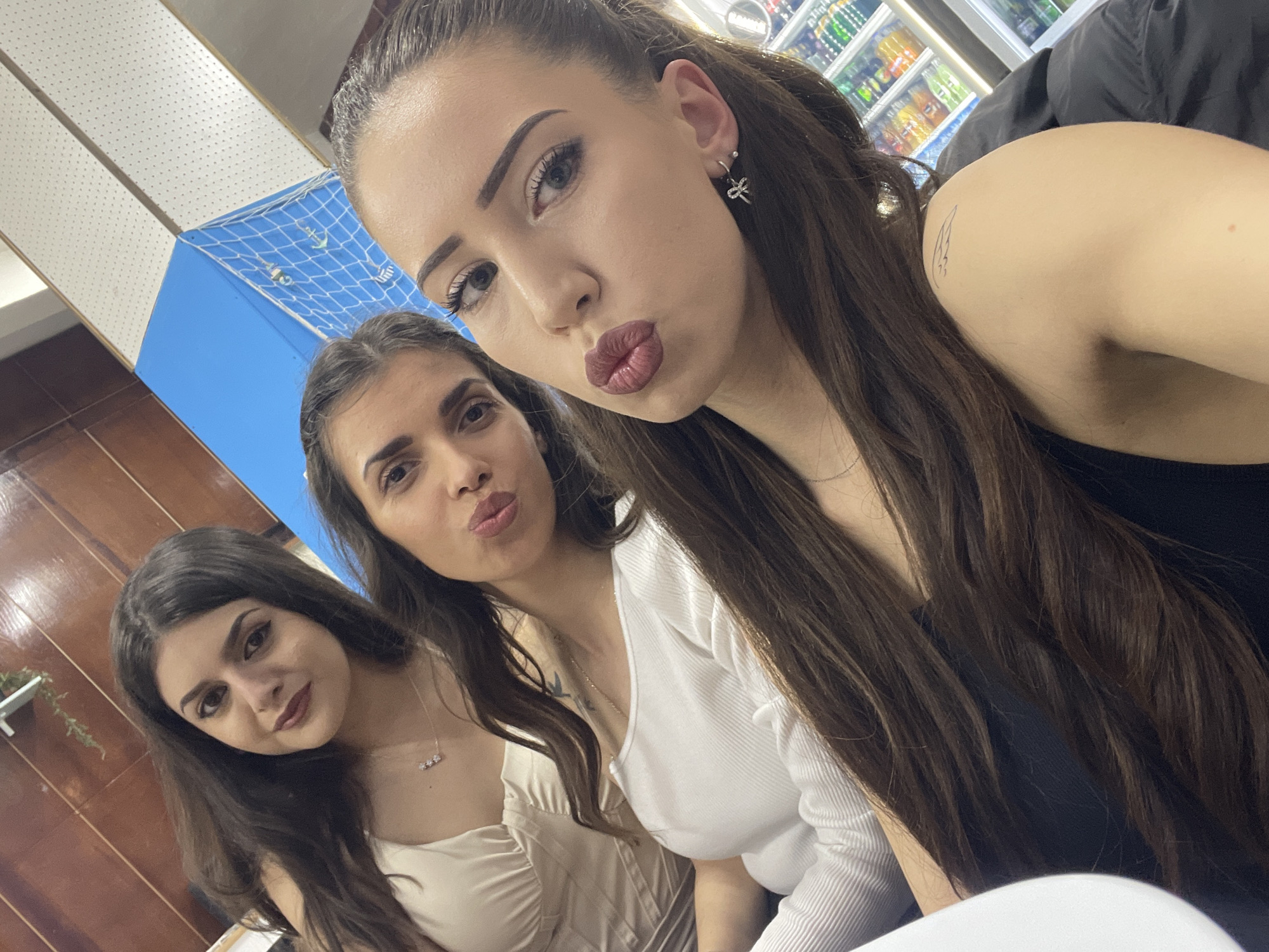 Three young women pose for a selfie. The woman closest to the camera has long, dark brown hair pulled back in a half ponytail, is wearing a black top and has a small tattoo of a bird on her upper right arm. Her hair is styled, and she is wearing a silver bow earring in her left ear and makeup. She is puckering her lips.
The second woman, in the middle, has dark brown hair and is wearing a white ribbed top. She is also puckering her lips. She has a tattoo on her chest.
The third woman, on the left, has shoulder length dark brown hair and is wearing a light colored top with a low neckline. She is wearing a necklace with charms.
Behind the women is a blue wall with a white net and a wall with wooden paneling. In the upper right corner of the image is a refrigerated display case stocked with beverages. The location is most likely in a shop. The city, Somovit, Bulgaria, cannot be definitively determined.