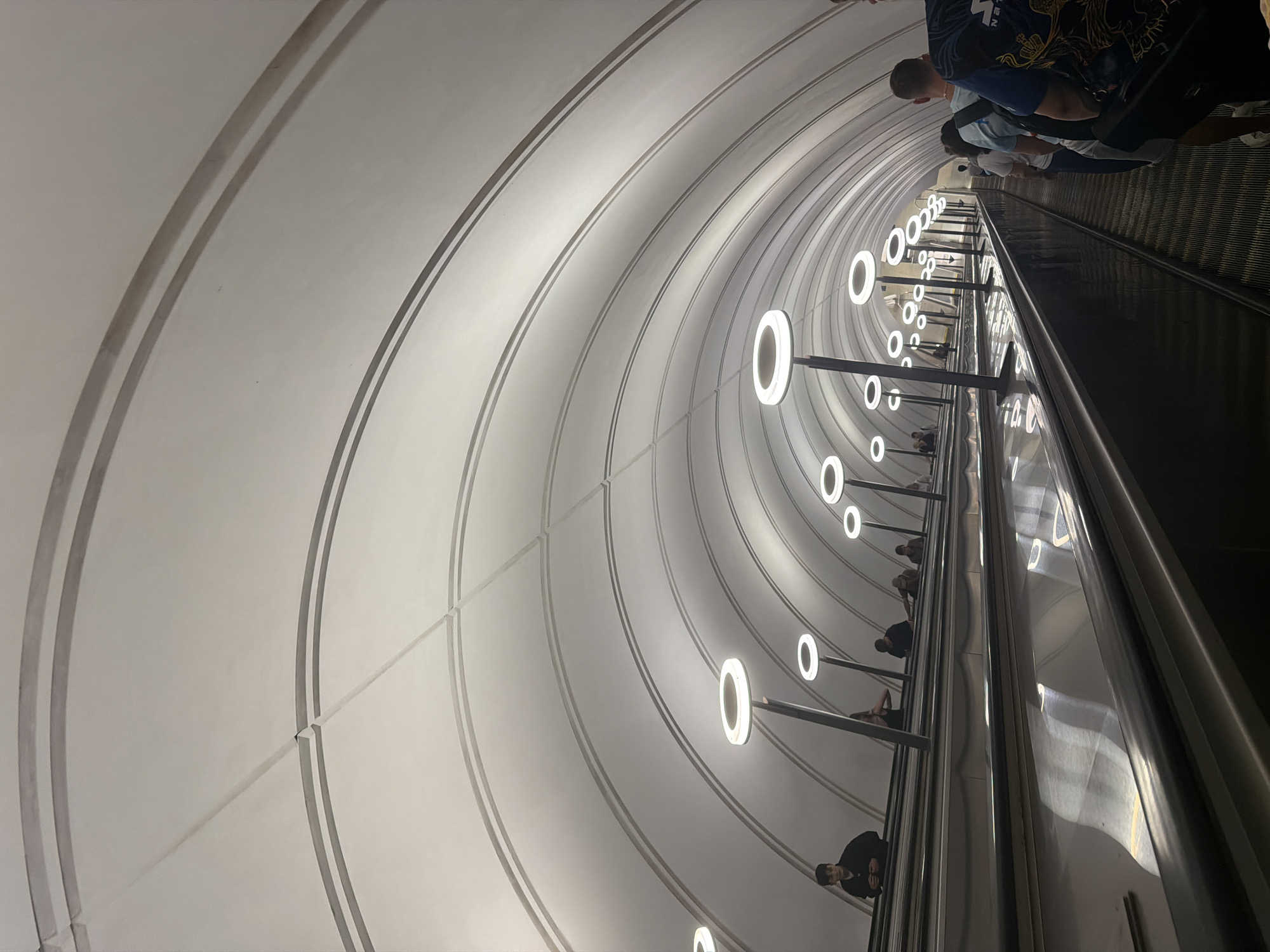 The image shows an interior view of a subway station, likely an escalator tunnel due to the presence of an escalator and multiple people ascending it. The setting is characterized by a large, curved, white ceiling with concentric ribbed detailing, illuminated by numerous circular, modern light fixtures that hang from dark poles. The escalator itself is a prominent dark metallic structure, with a continuous moving handrail and ribbed steps.
Several individuals are visible on the escalator, appearing to be heading upwards. They are dressed in casual clothing, suggesting a normal day's commute or travel. The lighting is bright and artificial, typical of underground public spaces, casting reflections on the metallic surfaces. The overall impression is one of architectural modernity and efficient public transport. There are no explicit indicators of time of day or weather, but the artificial lighting suggests it could be anytime, including nighttime. The location is not specified, but the style of the architecture and lighting is common in many contemporary metro systems. No visible text is present in the image.