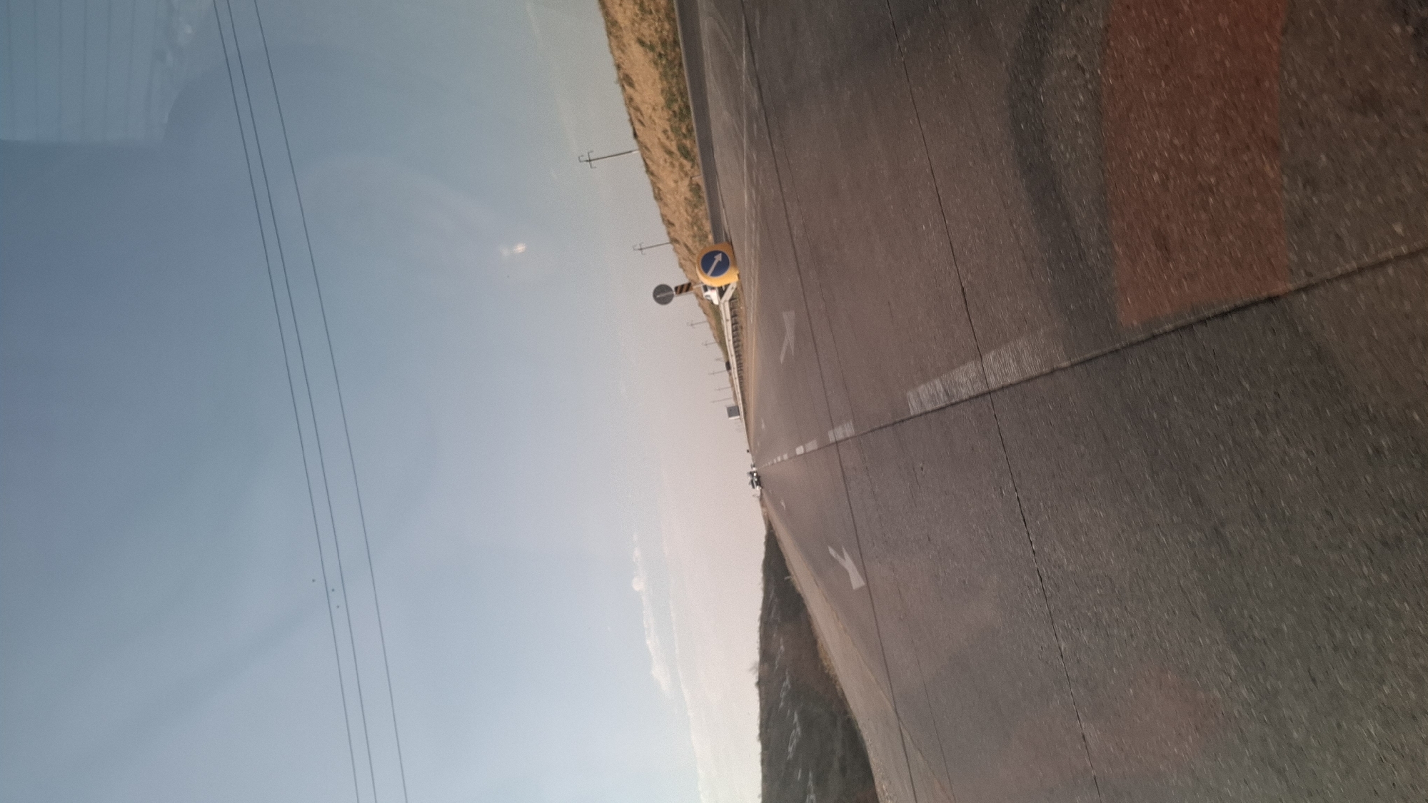An overpass or bridge with a highway is depicted. The road is paved and has lane markings. In the distance, there are vehicles. There is a traffic cone on the side of the road. The sky is clear and blue. There are also power lines visible. The location is Shymkent, Kazakhstan.