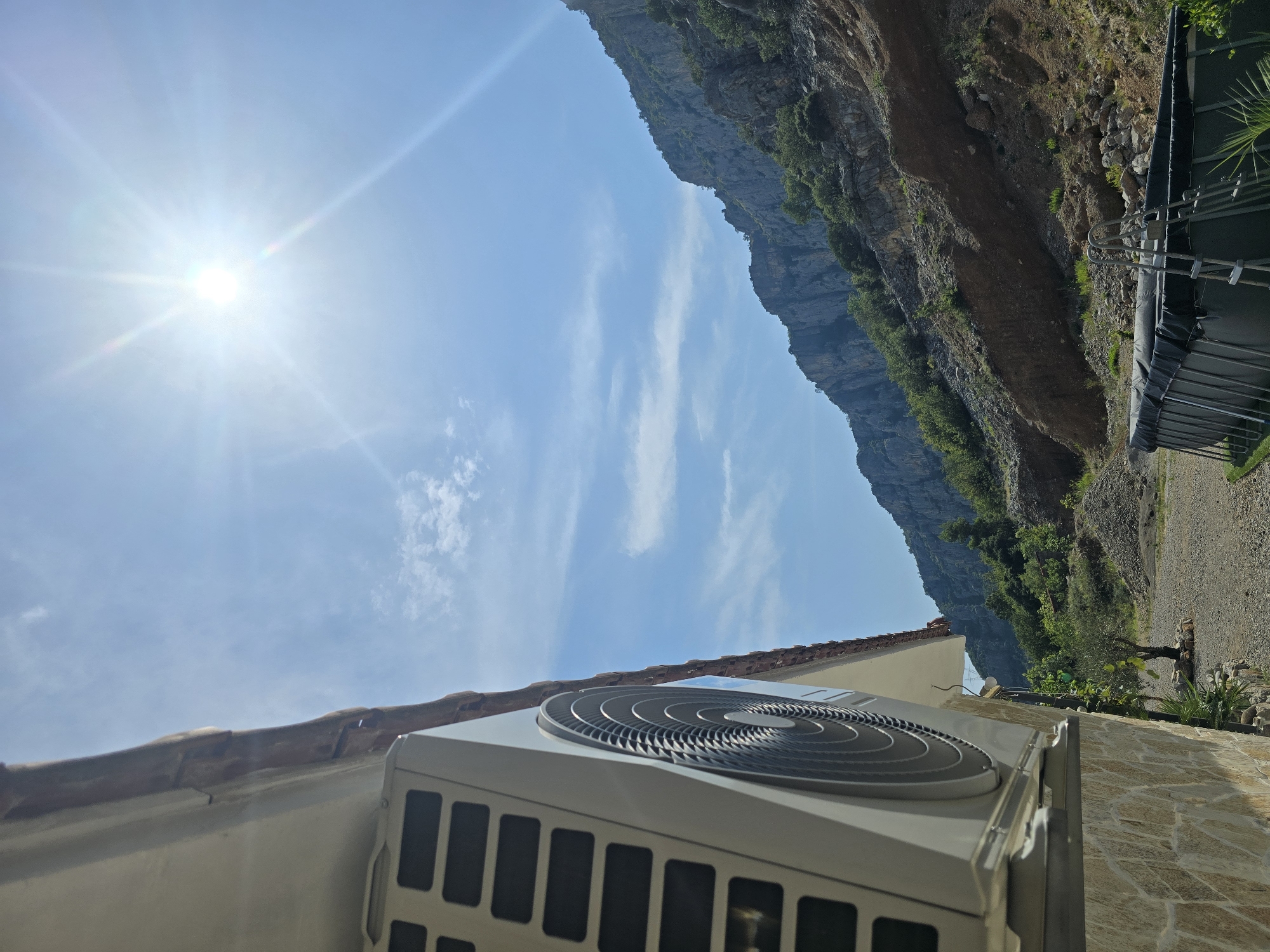 The image shows an outdoor scene on a sunny day. In the foreground, the side of a building with a stone facade and a white exterior unit of an air conditioner is visible. The air conditioner unit has a fan with circular fins and a grille. The building has a tiled roof overhang. To the right of the building, there is a gravel ground with some vegetation. Further in the background, there is a large, dark-colored swimming pool with a ladder. Beyond the pool, the terrain rises steeply, forming a rocky mountainside covered with sparse green vegetation. The sky is blue with some wispy white clouds, and the sun is shining brightly, creating lens flare. The scene appears to be in a natural, possibly rural or mountainous, setting.
