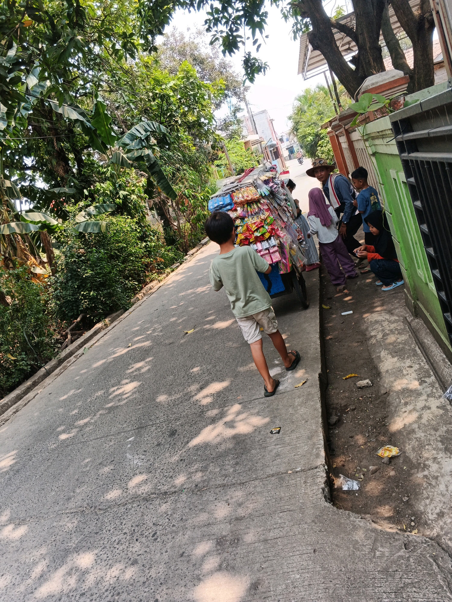 In Kadu, Indonesia, on a sunny day, a boy in a green t-shirt and shorts walks away from a cart laden with colorful snacks. He is on a paved road lined with lush green trees and vegetation. To the right of the road, a group of people are gathered near a fence. A man in a hat, a woman in a purple headscarf, two boys, and a girl in a black headscarf are visible. The girl is crouching and looking at something in her hands. The scene suggests a casual, everyday street life.