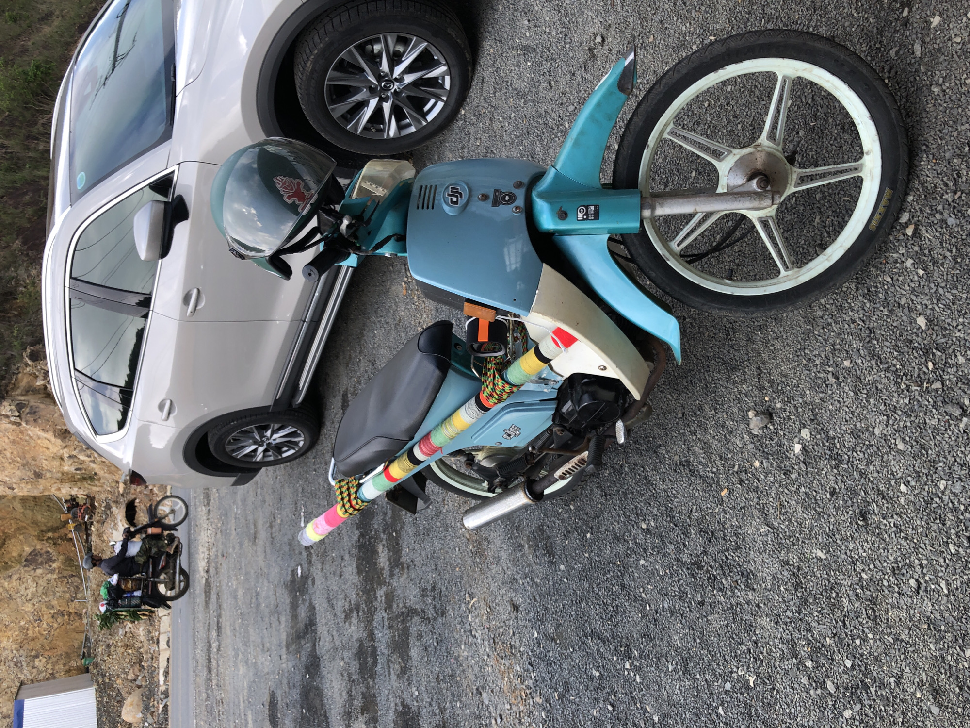 In Nam Hải, Vietnam, a light-colored SUV is parked on a gravel lot. In front of the SUV, a light blue moped is parked. The moped has a teal-colored body and a black seat. A green helmet with a red emblem is placed on the handlebars. The rear rack of the moped is decorated with colorful yarn wrapping. A second motorcycle, ridden by a person wearing a helmet and camouflage pants, is visible in the background, loaded with bundles of greenery. The environment consists of a rocky hillside and a paved road.