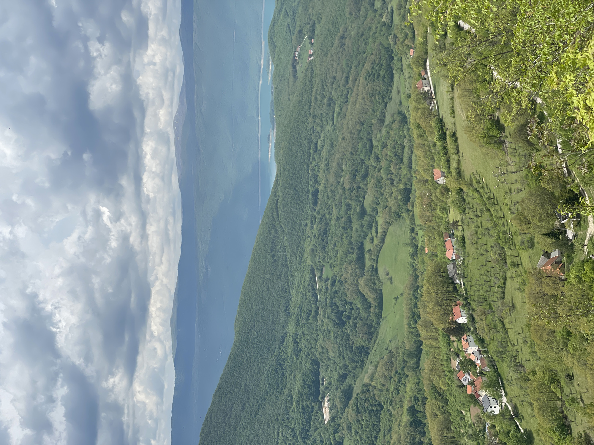 The image shows a panoramic view from a high vantage point in Gmići, Bosnia and Herzegovina. The landscape features a deep blue lake or river alongside a dense green forested mountain range. The sky is filled with billowing gray and white clouds. A rural area with scattered houses having red roofs is visible along a sloping green hillside. Some trees and shrubs are in the foreground.